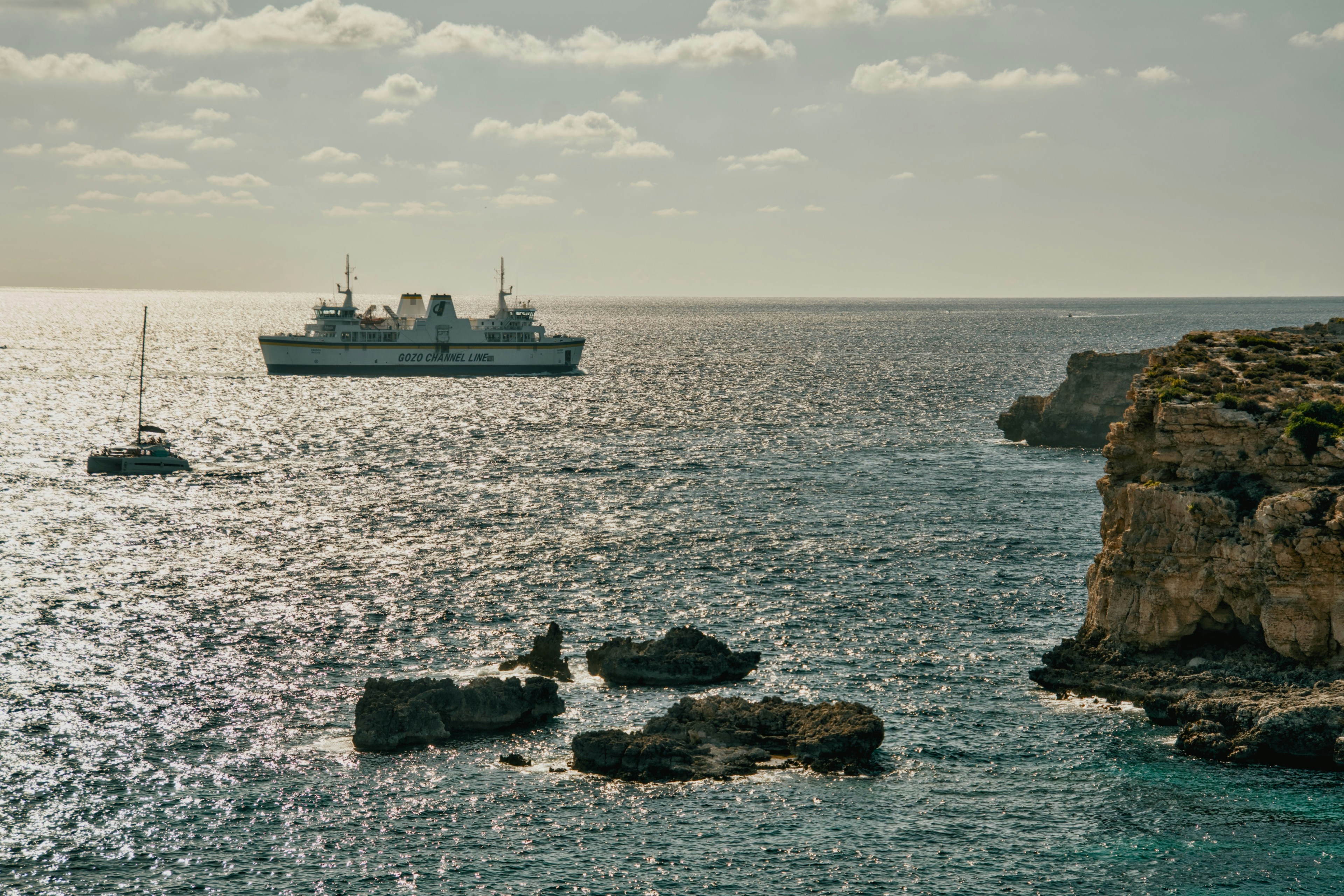 A Gozo Channel Line ferry and a sailboat on sparkling water near rocky cliffs