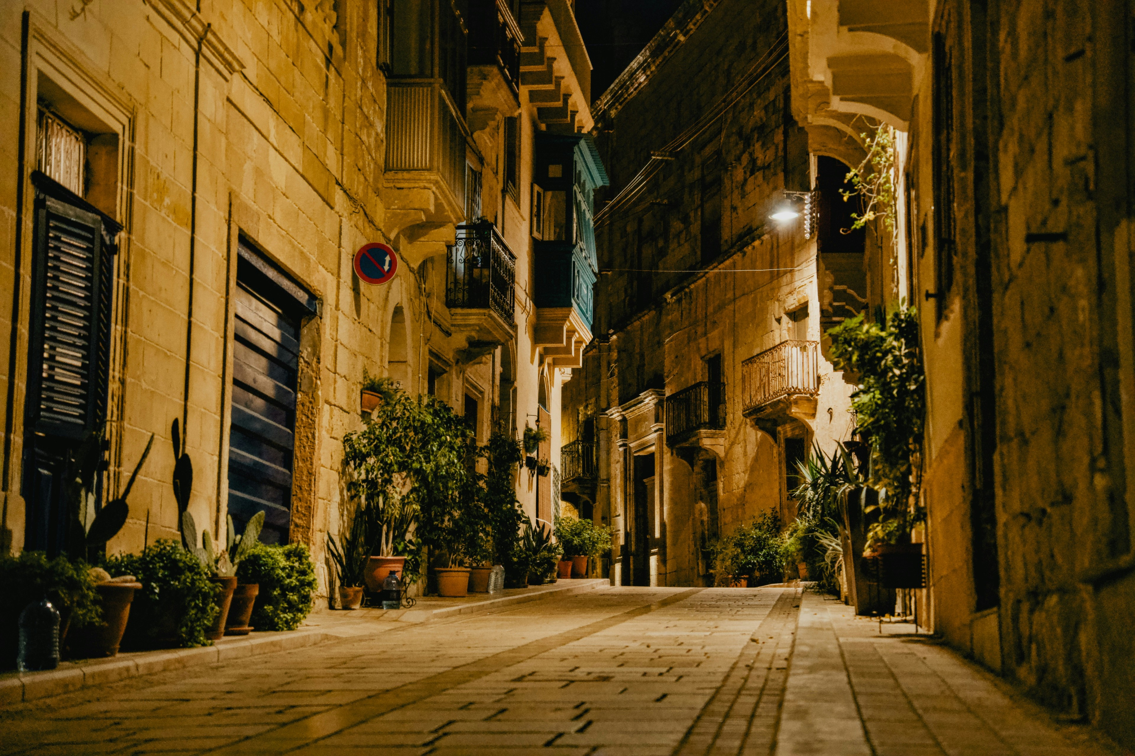 A narrow stone street at night with potted plants and warm yellow street lighting