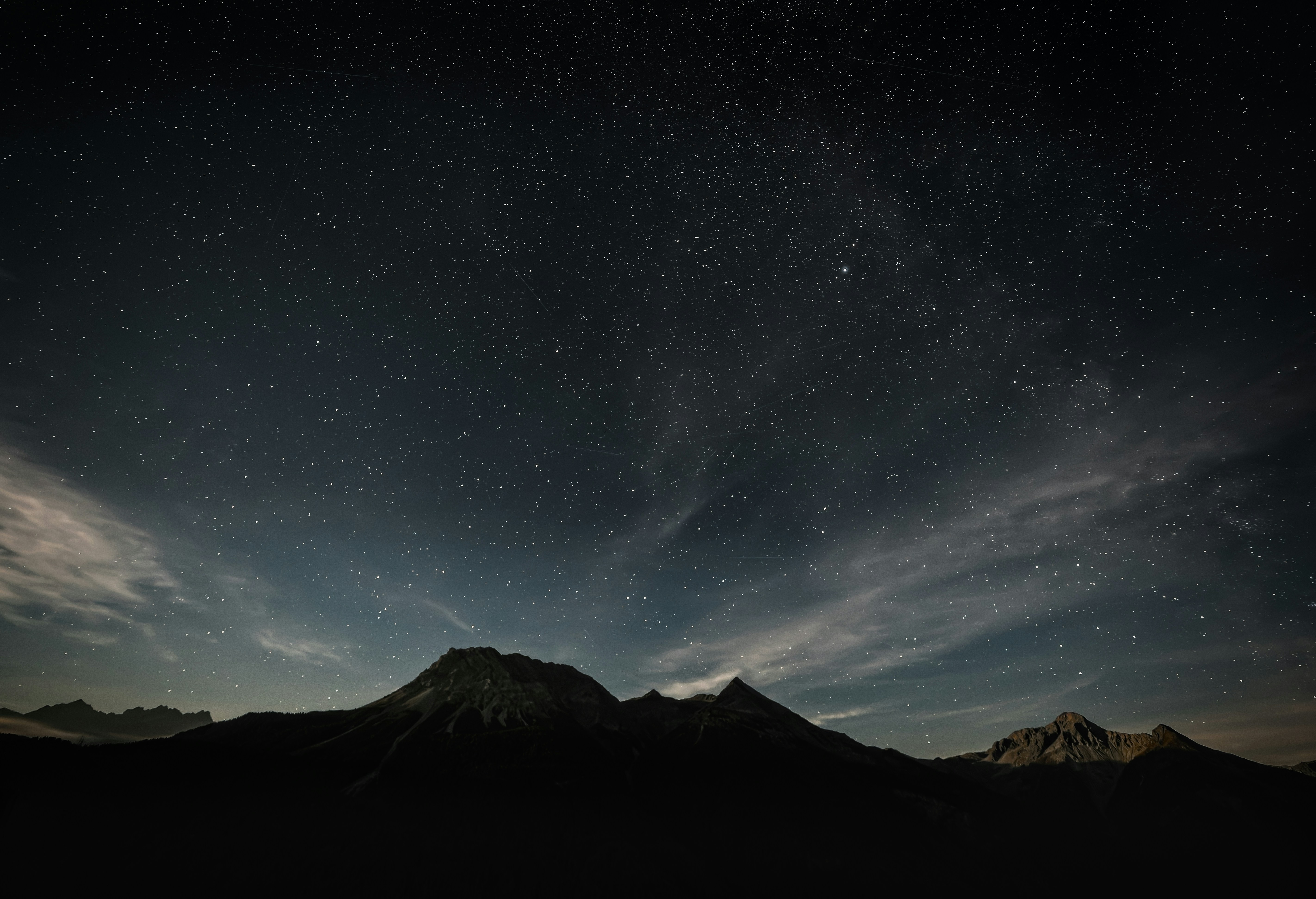 A starry night sky above dark mountain silhouettes and wispy clouds