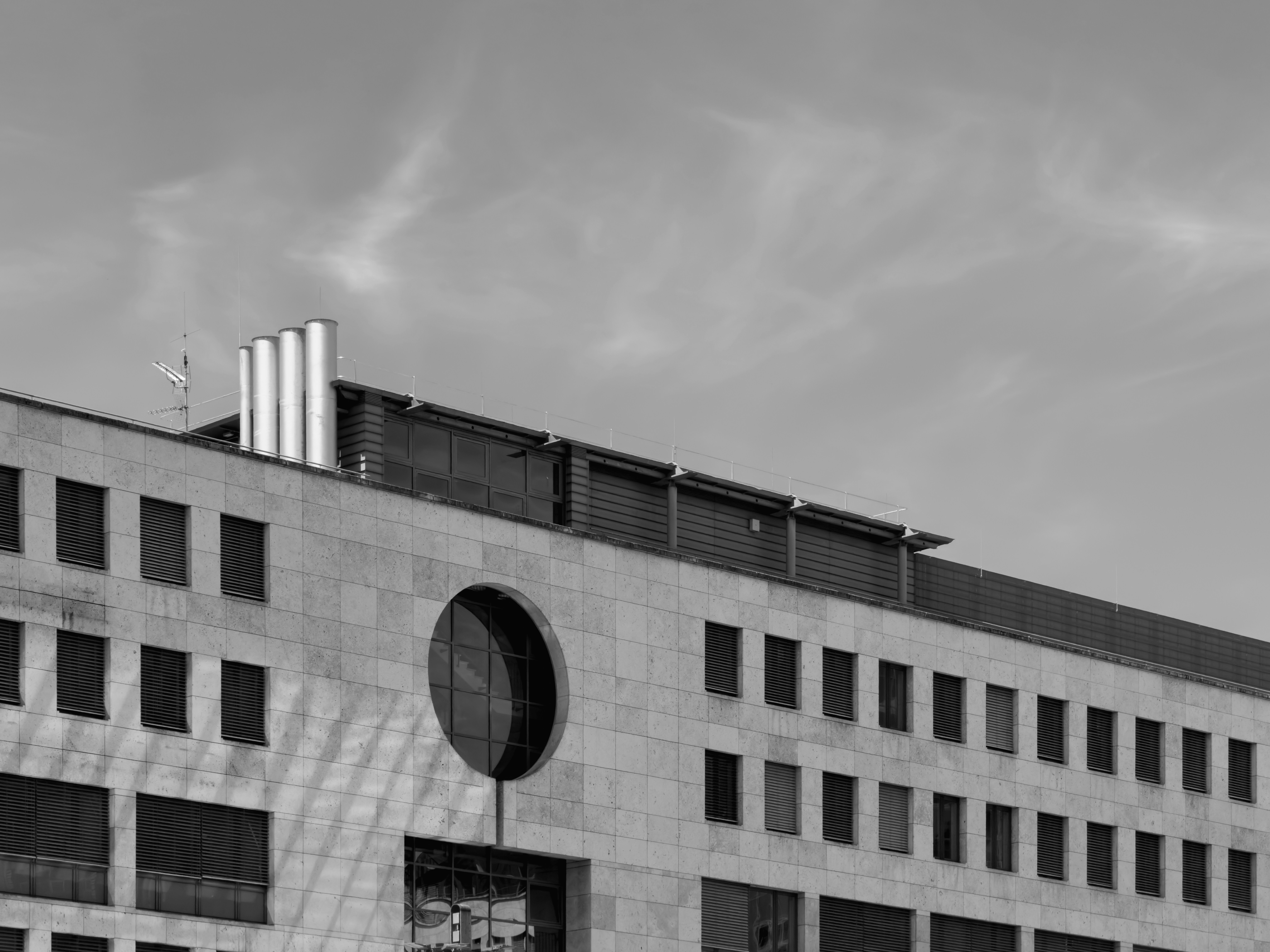 Eine steinerne Gebäudefassade mit einem runden Fenster und Metallschornsteinen unter einem wolkigen Himmel