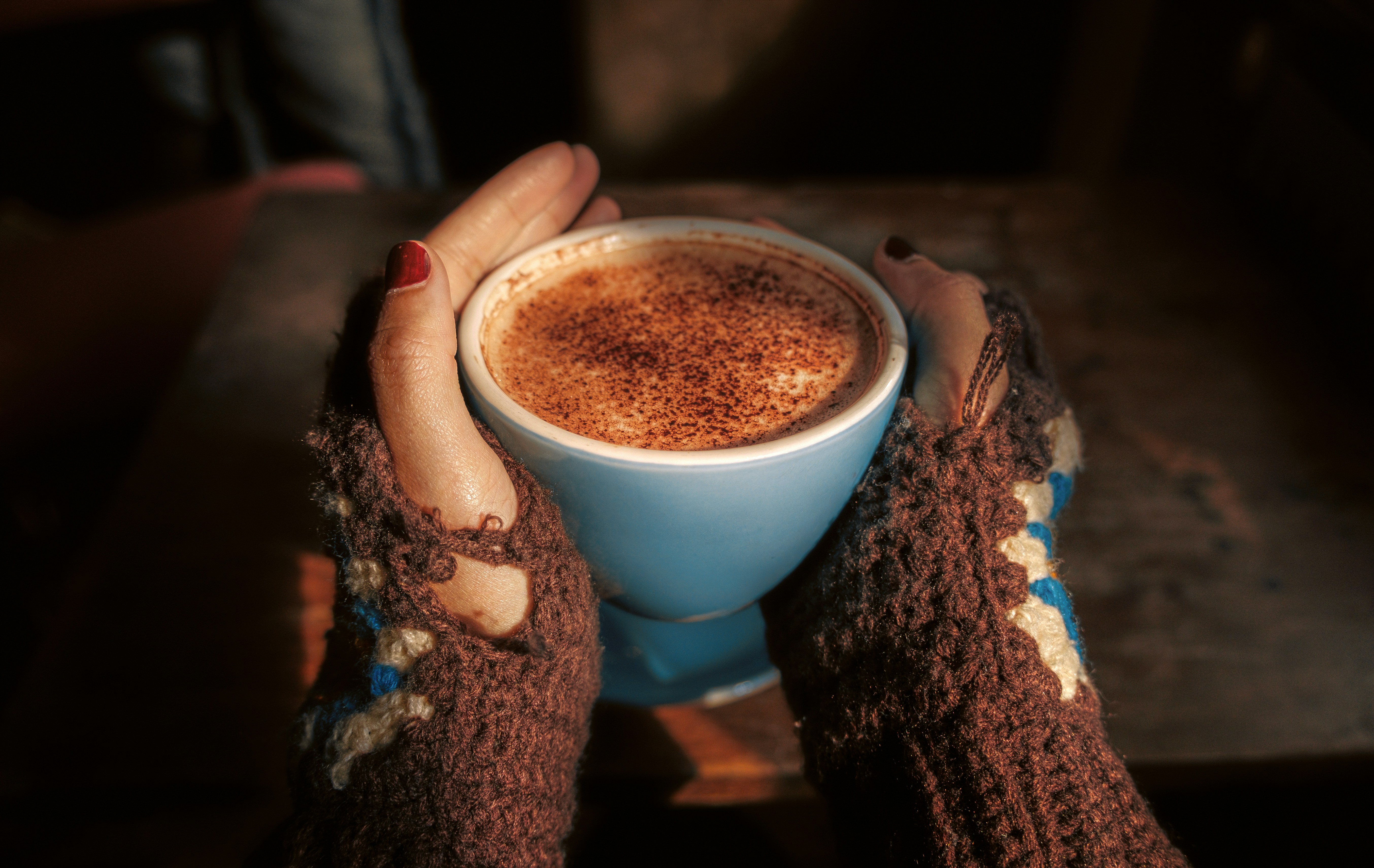Mani in guanti di maglia marroni senza dita che tengono una tazza blu di caffè