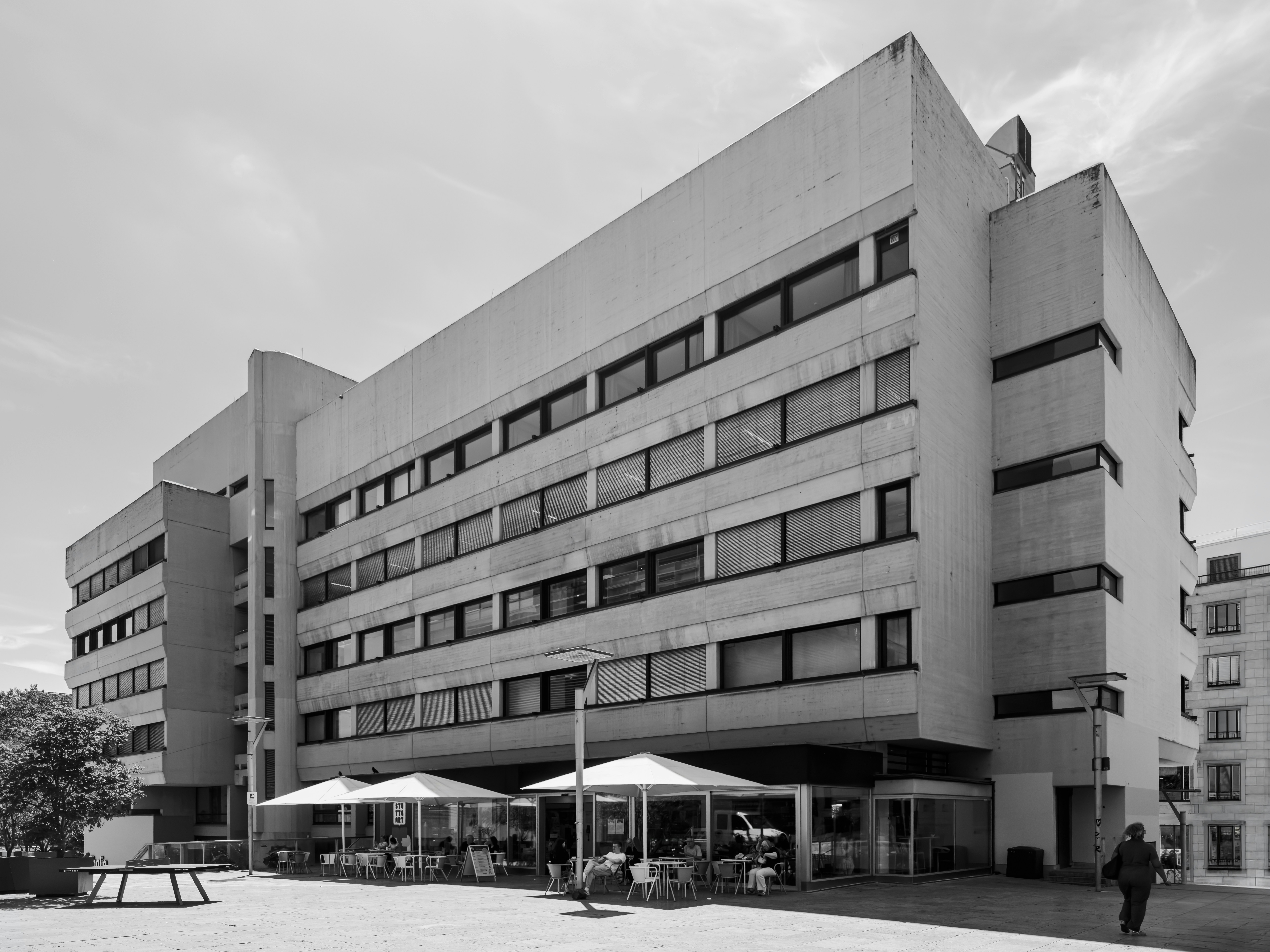Ein großes Bürogebäude aus Beton mit Außen-Cafés unter weißen Sonnenschirmen