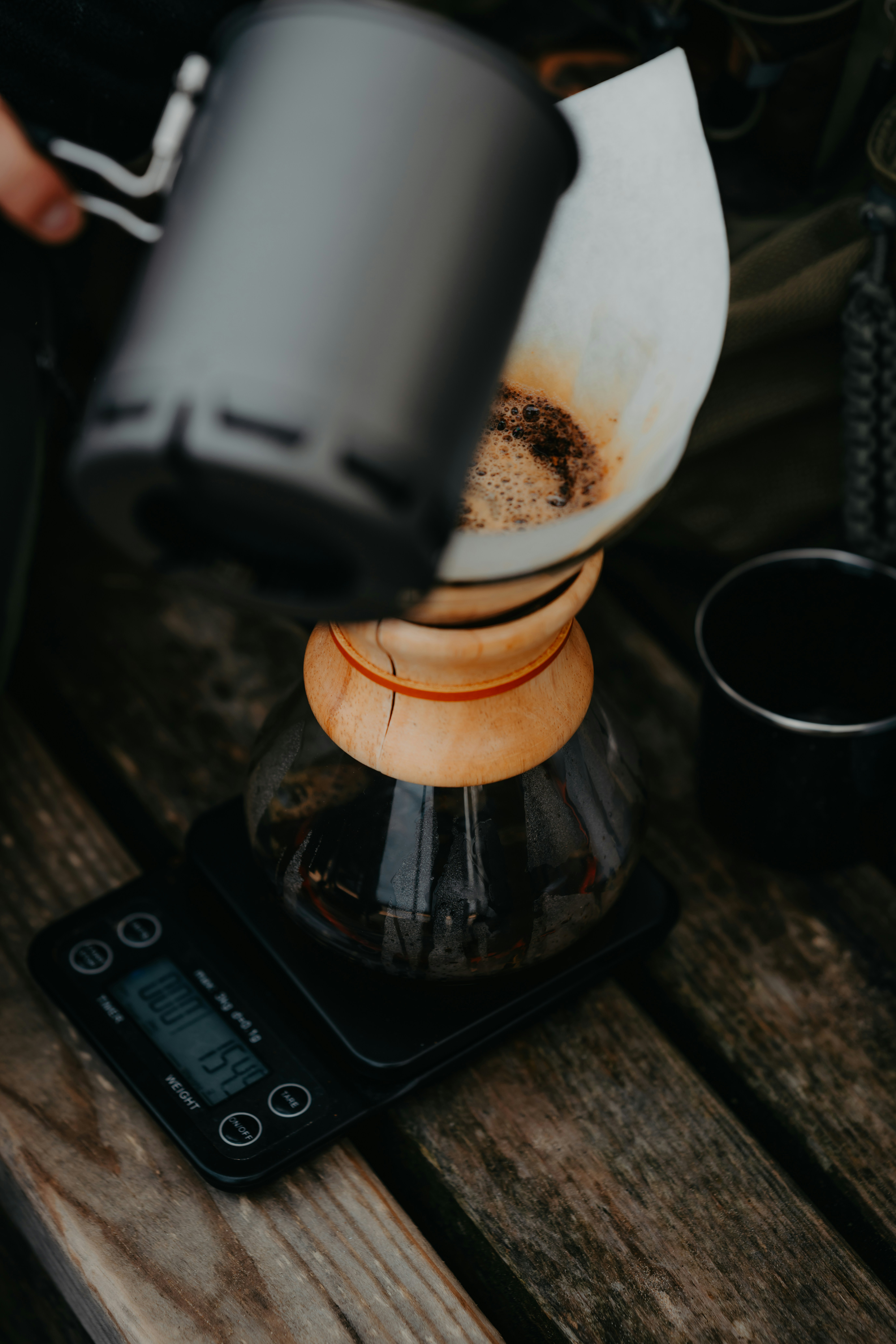 A person pouring hot water from a metal kettle into a Chemex coffee maker