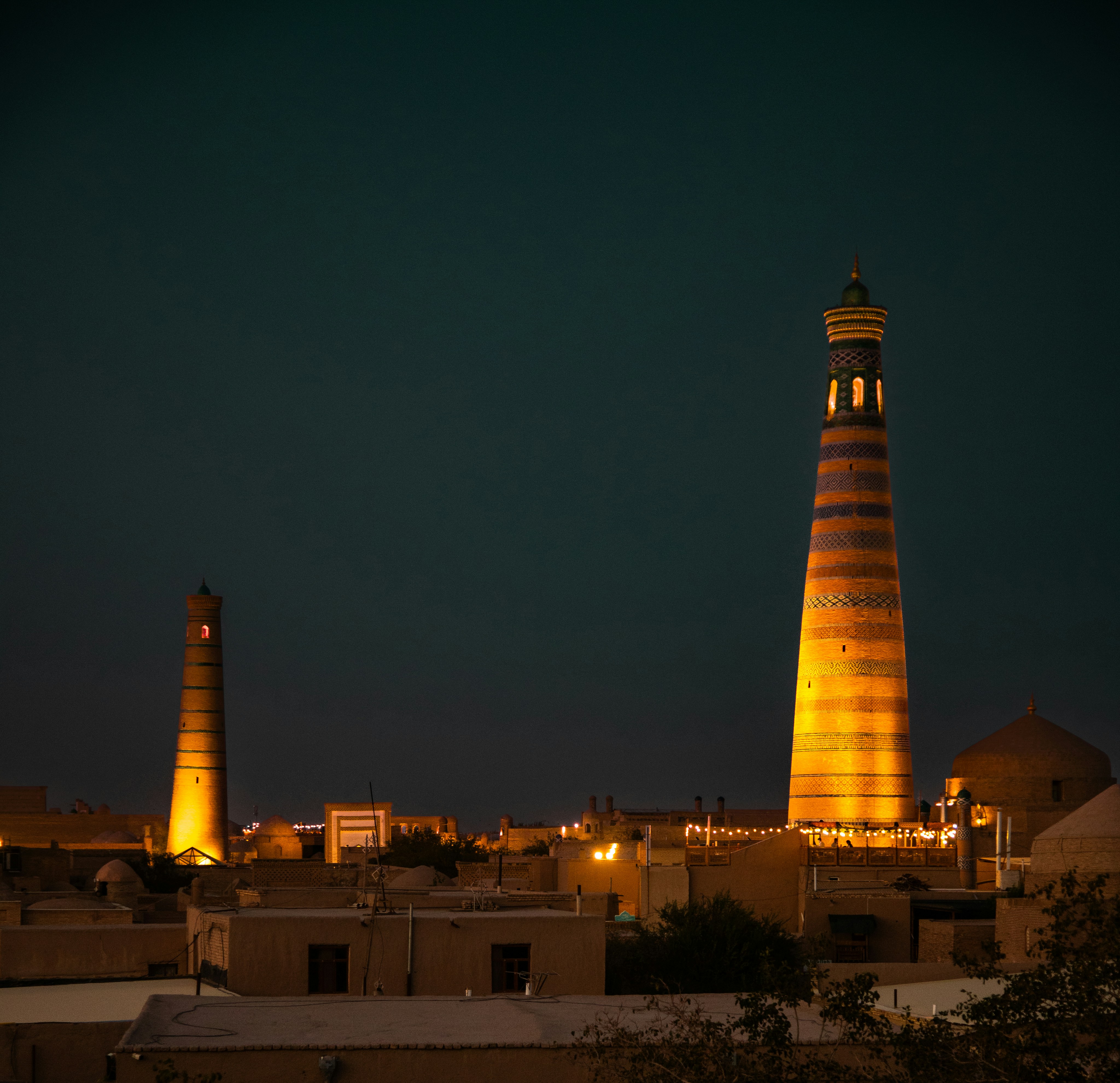 Dois minaretes iluminados se erguendo acima dos telhados planos de uma cidade antiga à noite