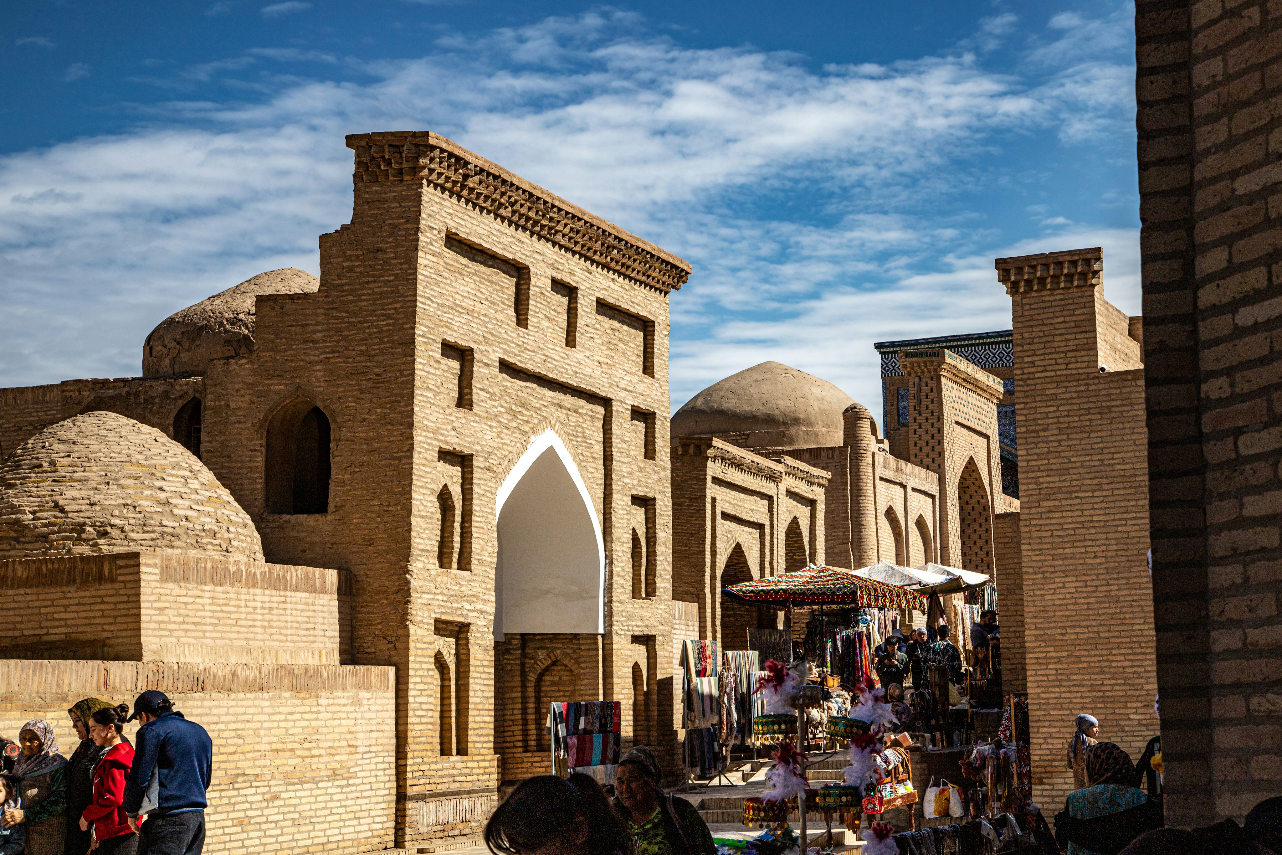 Pessoas passando por antigos edifícios de tijolos com portas em arco e cúpulas em Khiva