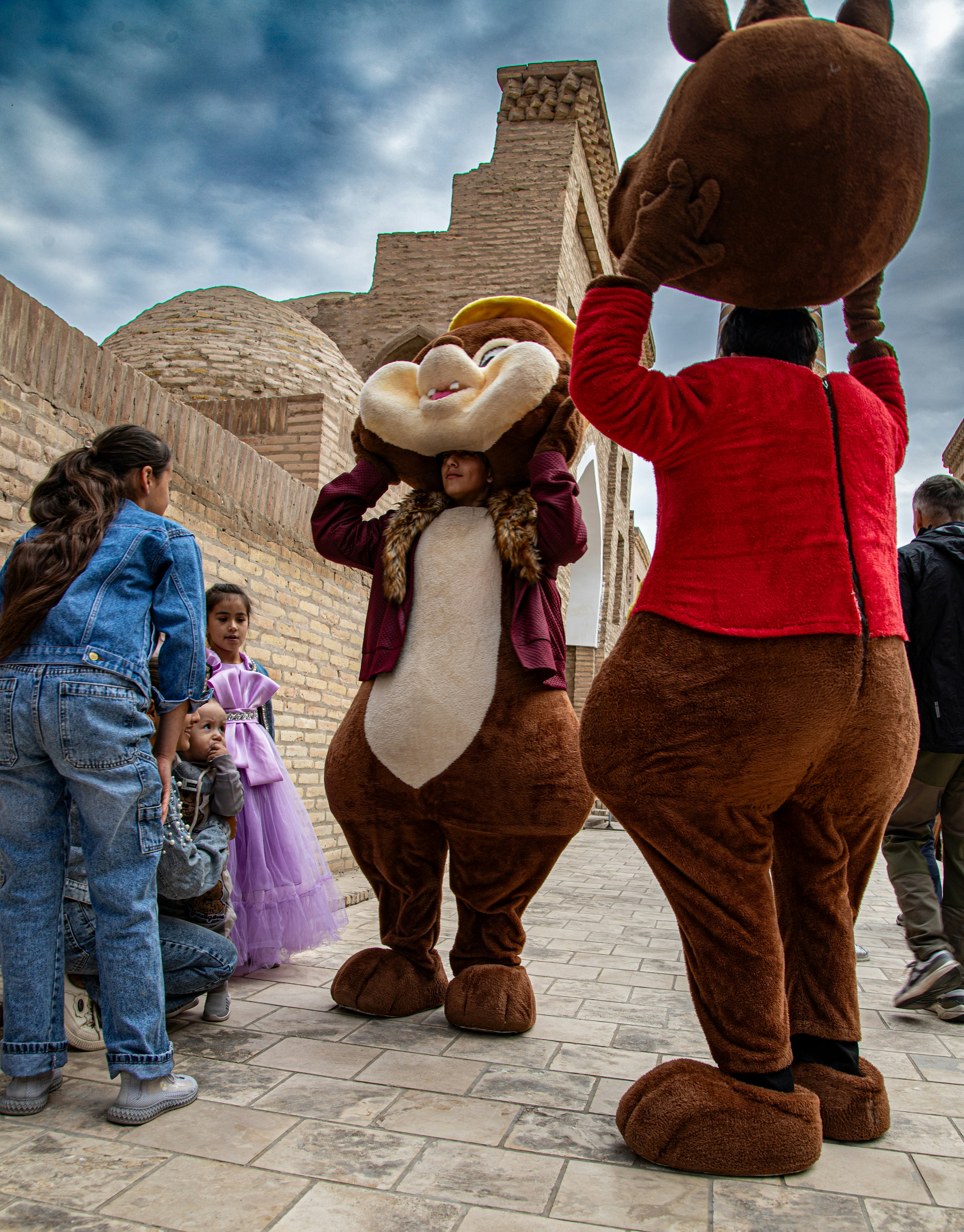 Duas pessoas com fantasias marrons de mascote animal interagindo com crianças perto de um prédio de tijolos