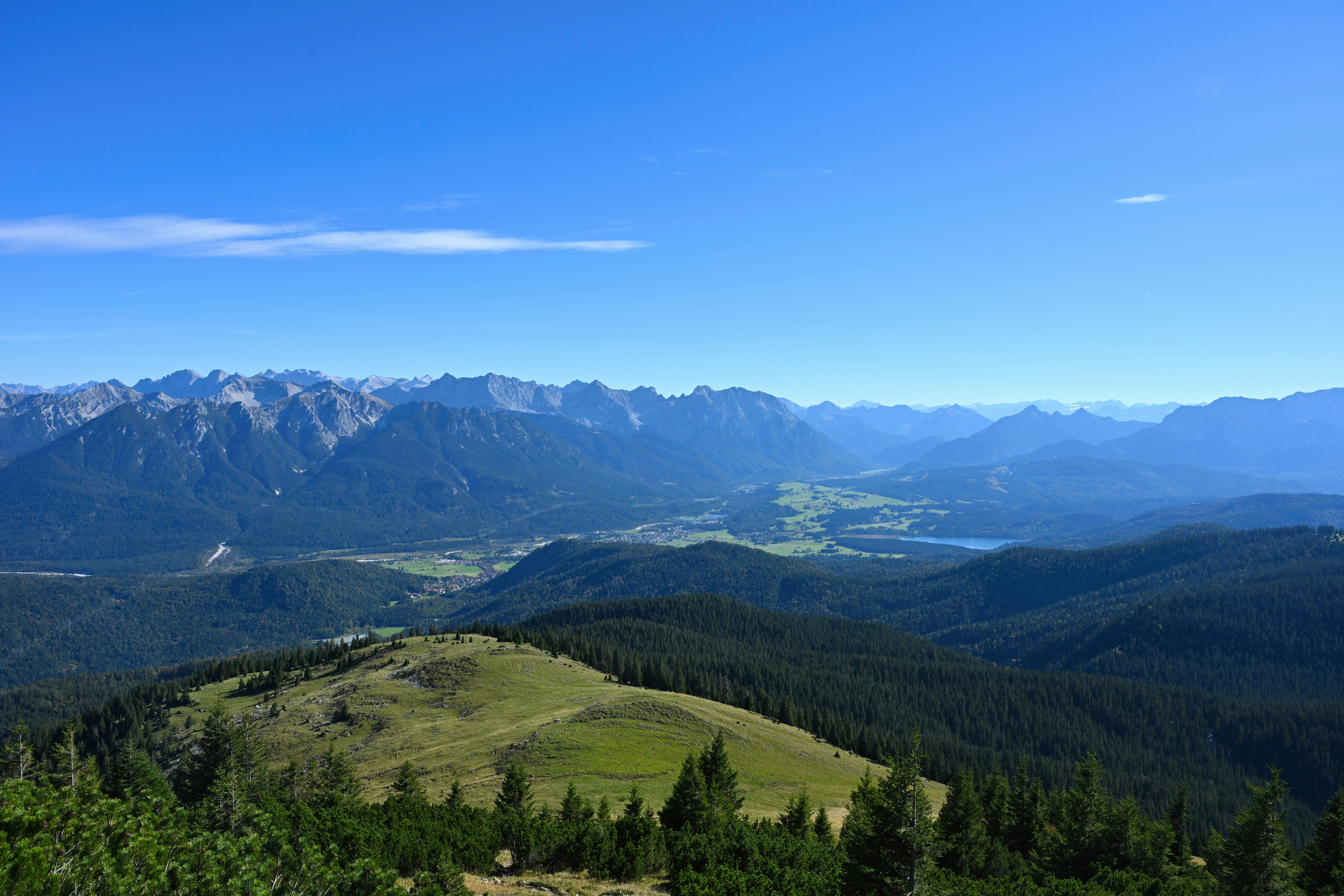 A green mountain slope overlooking a valley with a lake and distant peaks