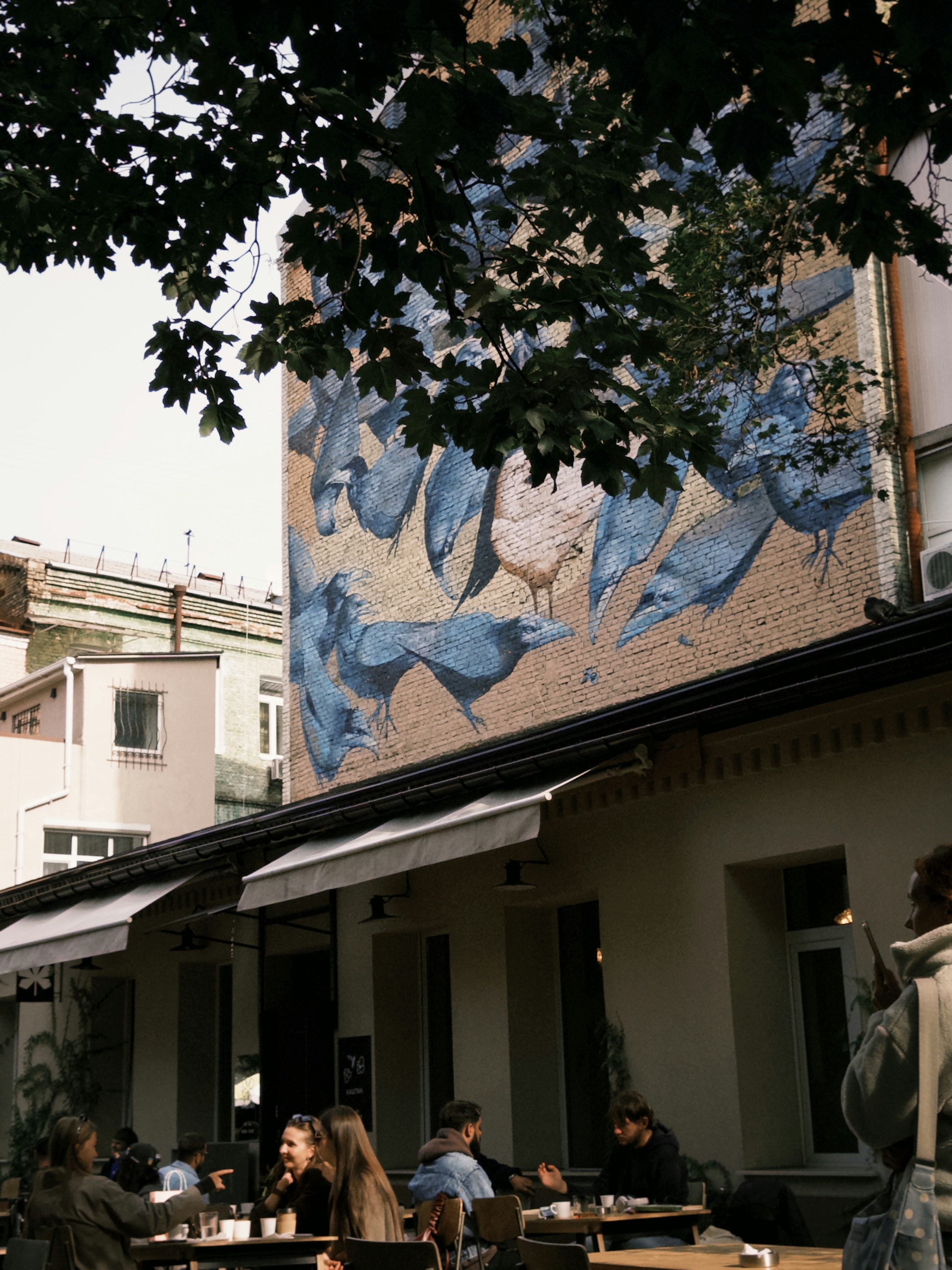 Pessoas sentadas em mesas ao ar livre sob uma parede de tijolos com um mural de pássaro azul