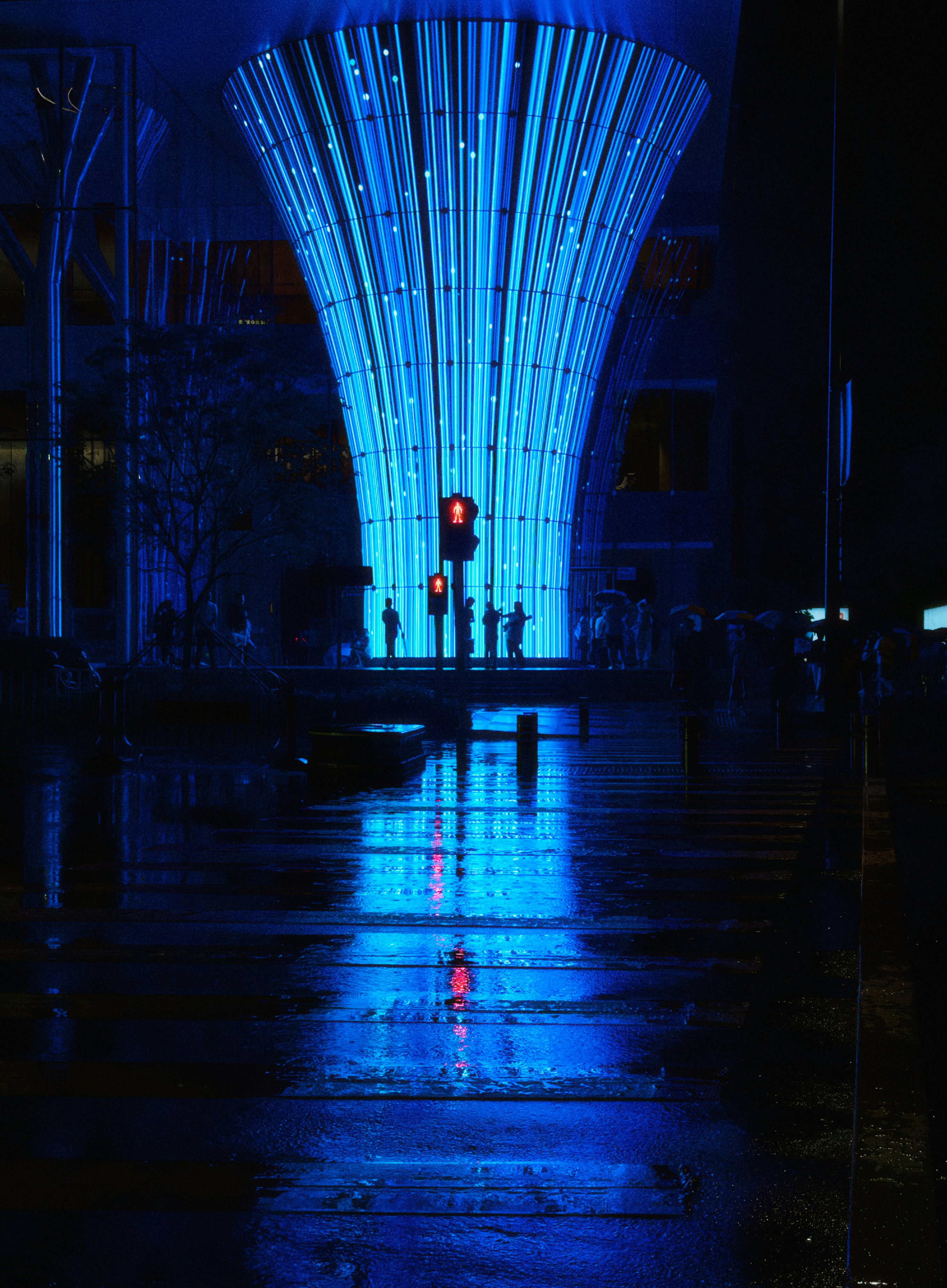 Silhuetas de pessoas atravessando uma rua molhada sob uma instalação de luz azul brilhante