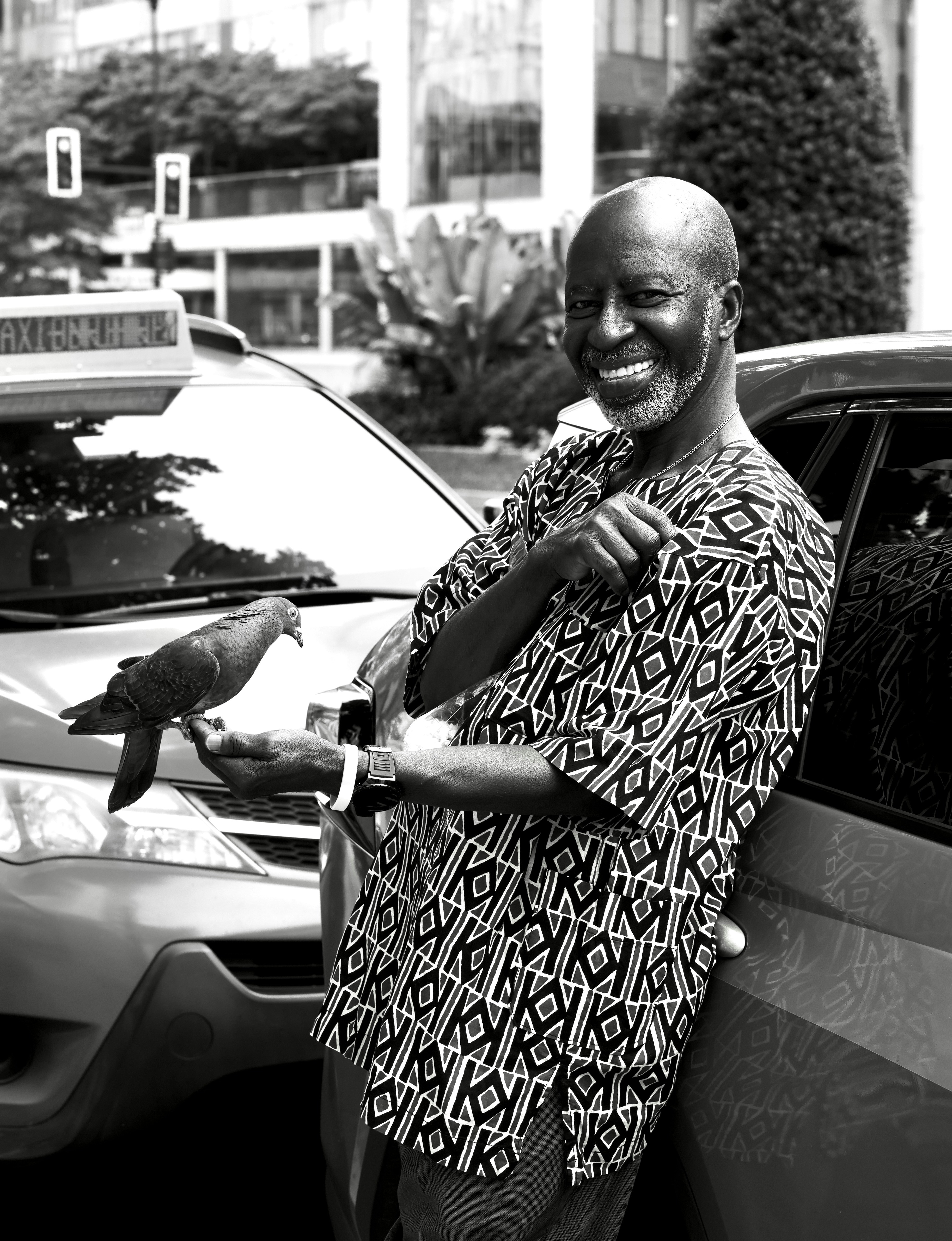 A smiling man in a patterned shirt holding a pigeon beside a taxi