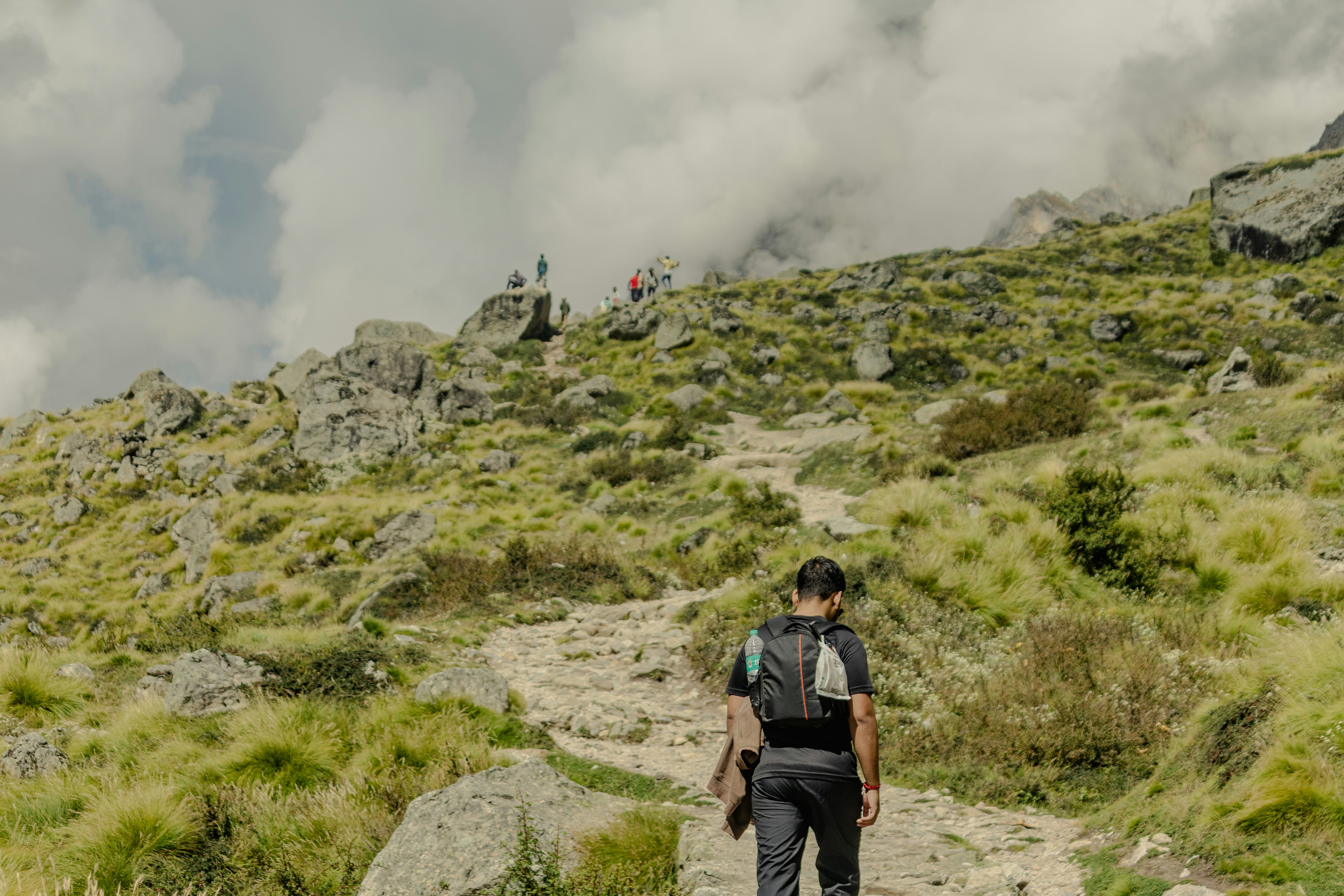 A person with a backpack hiking up a rocky mountain trail toward distant figures