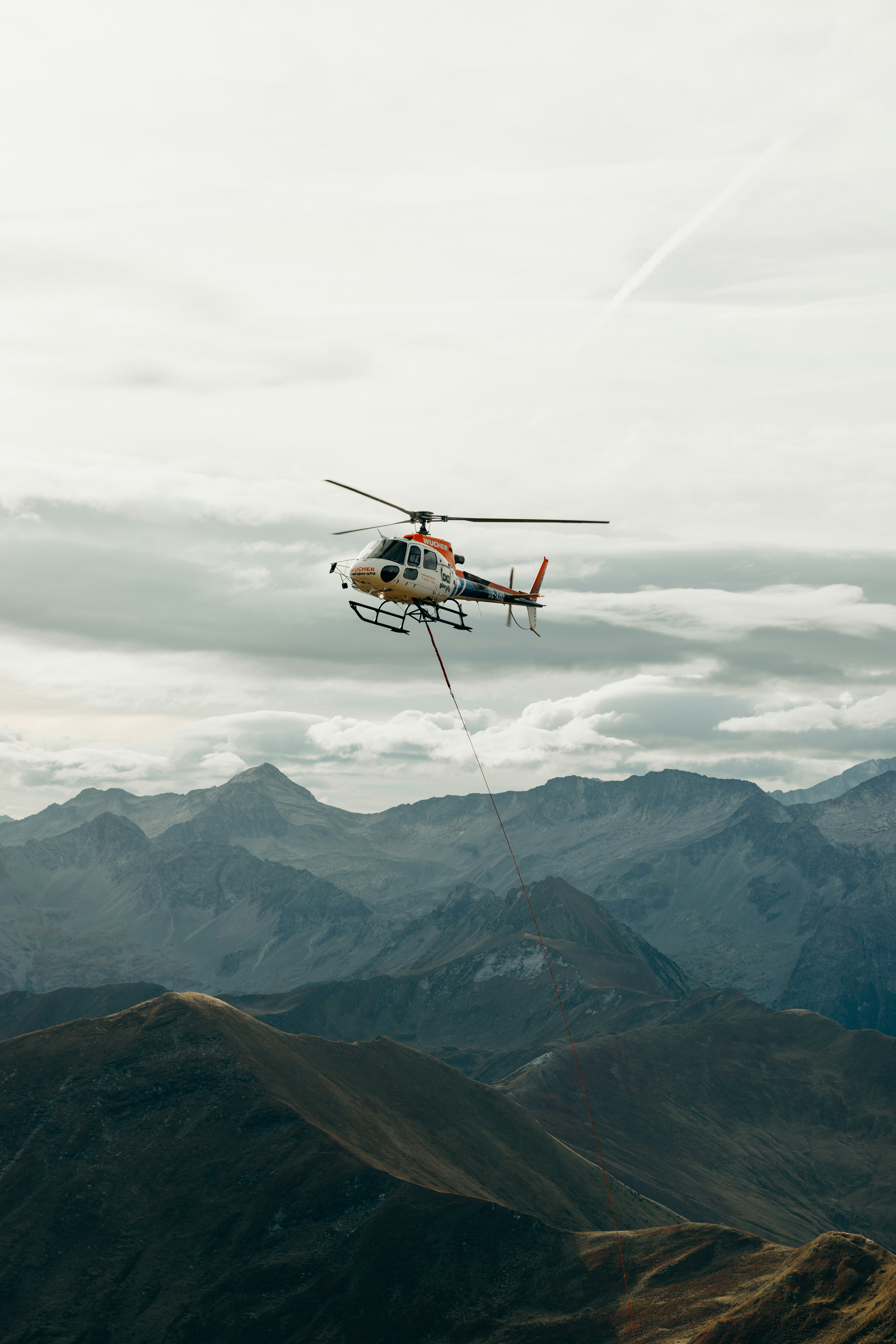 A helicopter with a long cable flying over rugged mountain peaks under a cloudy sky