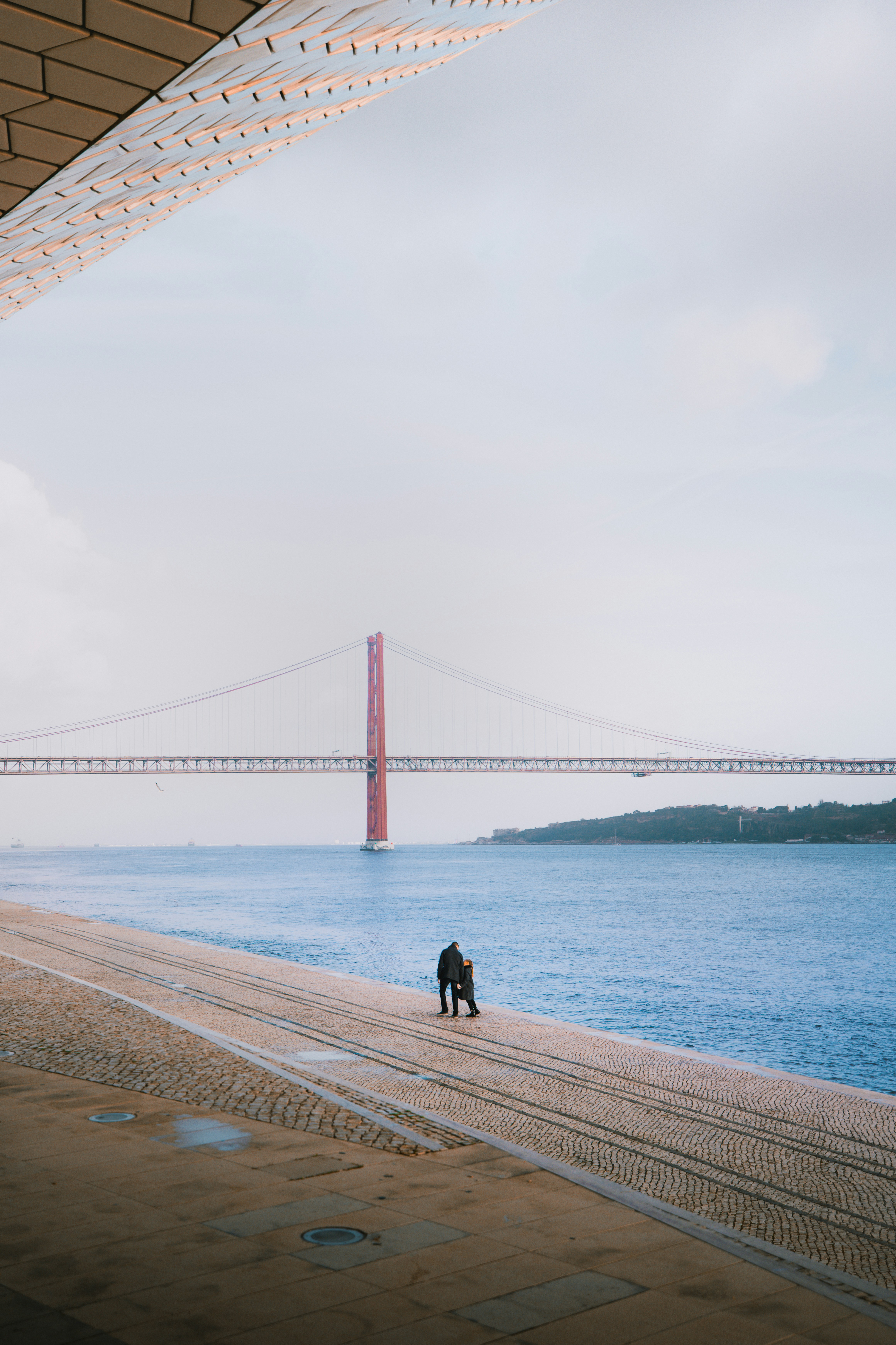 Due persone che camminano su una passeggiata sul lungomare vicino al Ponte 25 de Abril a Lisbona