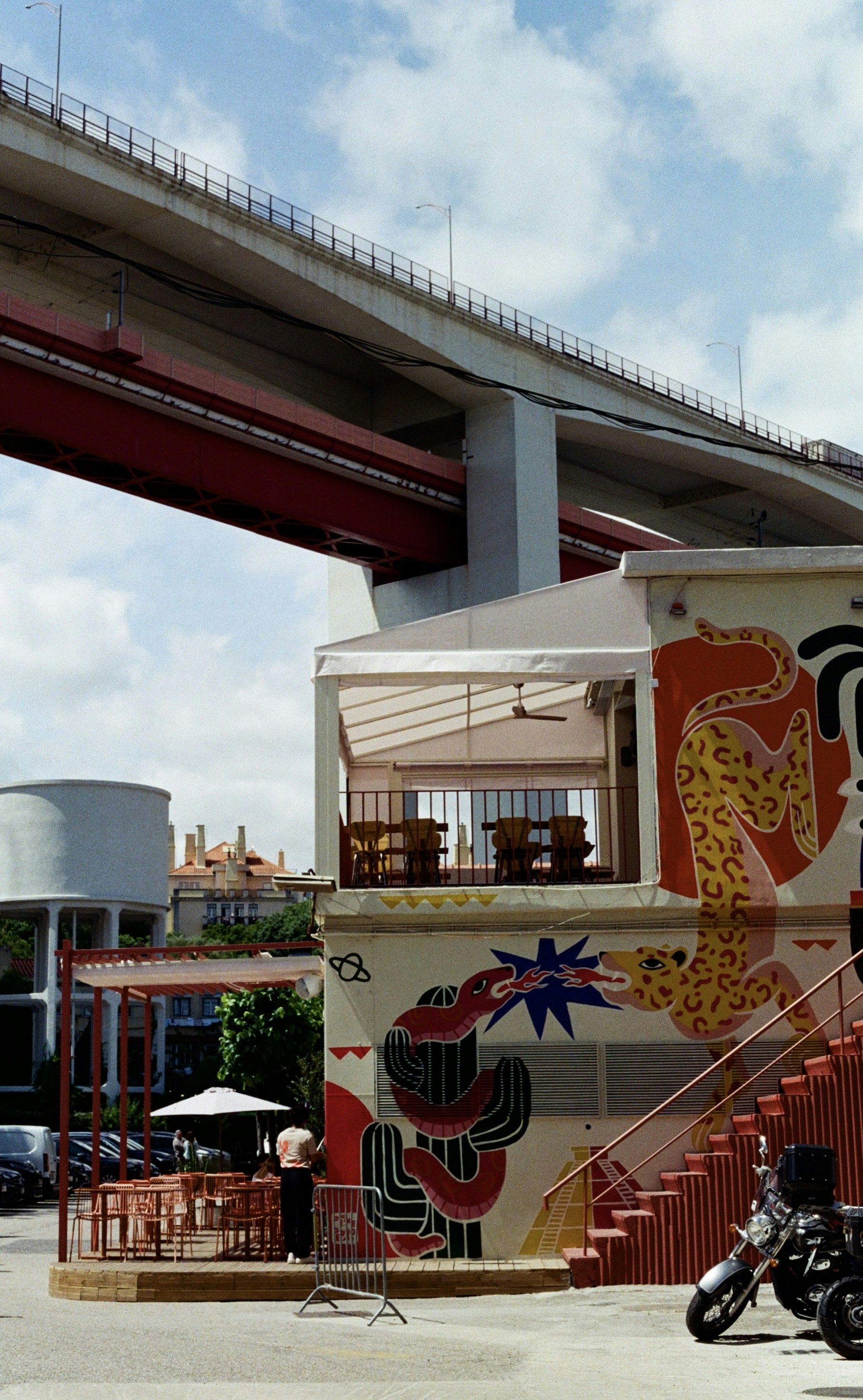 A building with colorful murals under a large red bridge on a sunny day