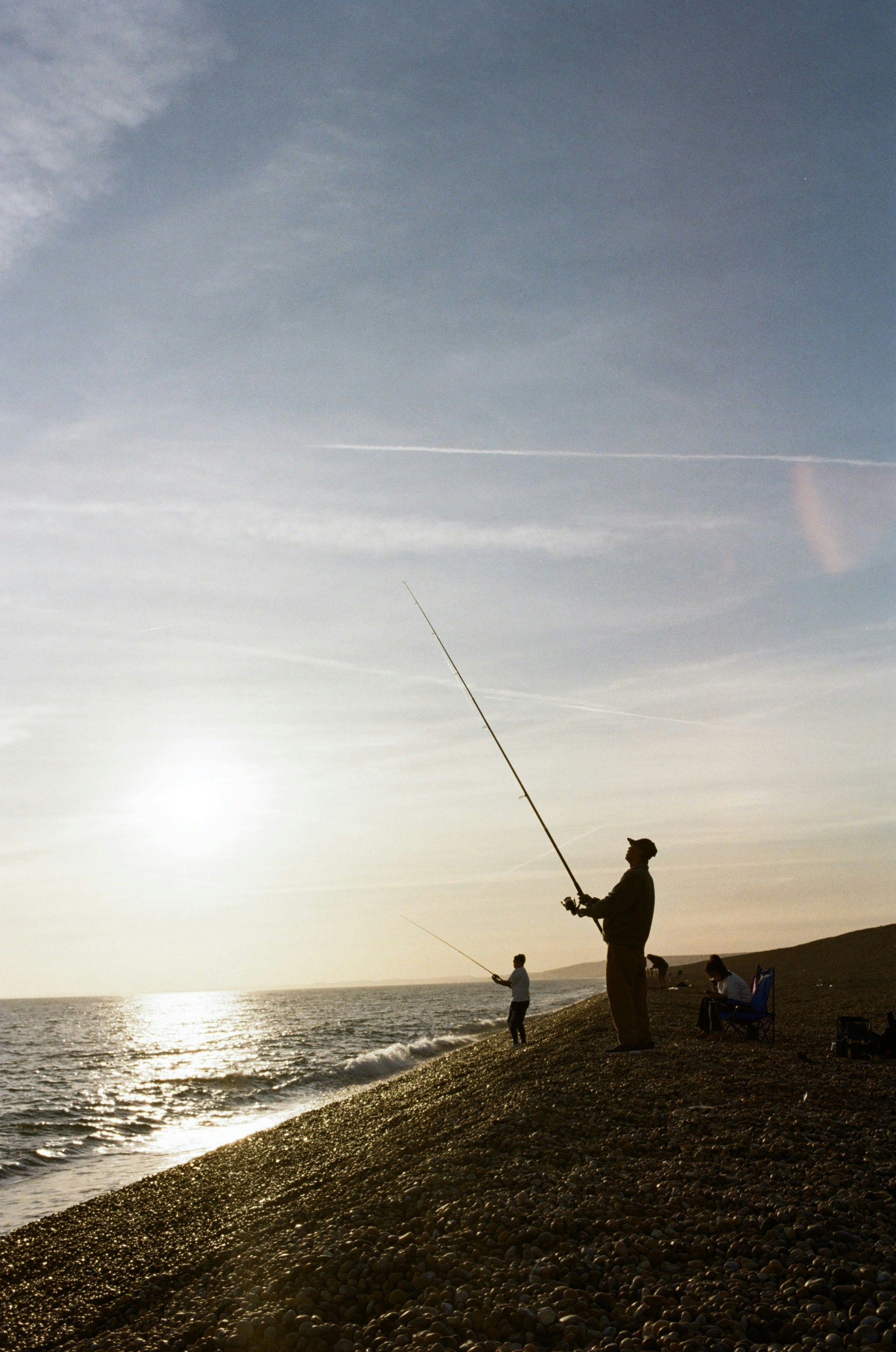 People fishing on a pebble beach under a bright low sun