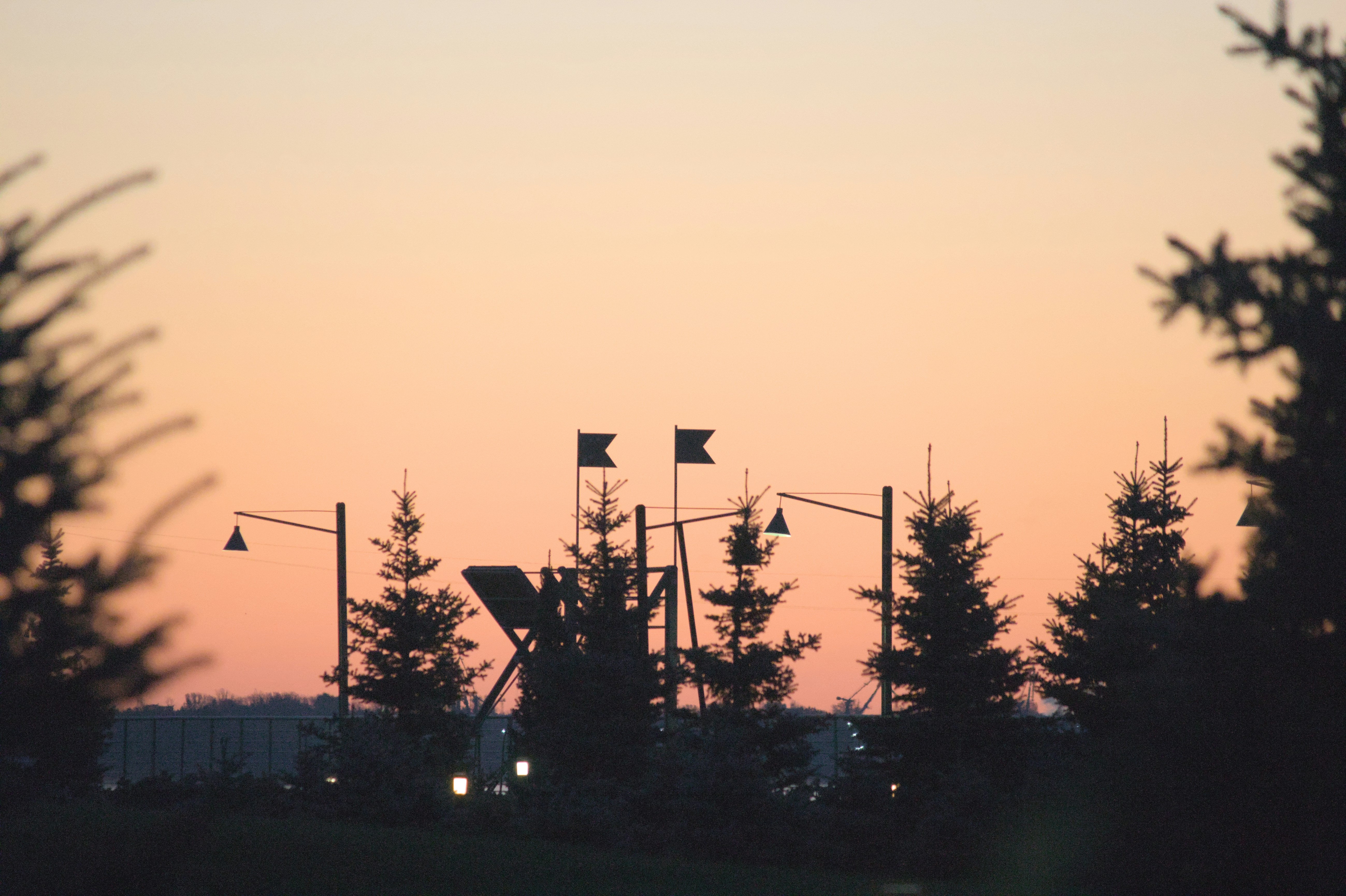 Silhouettes of fir trees, flags and lampposts stand out against a soft peach sunset sky.