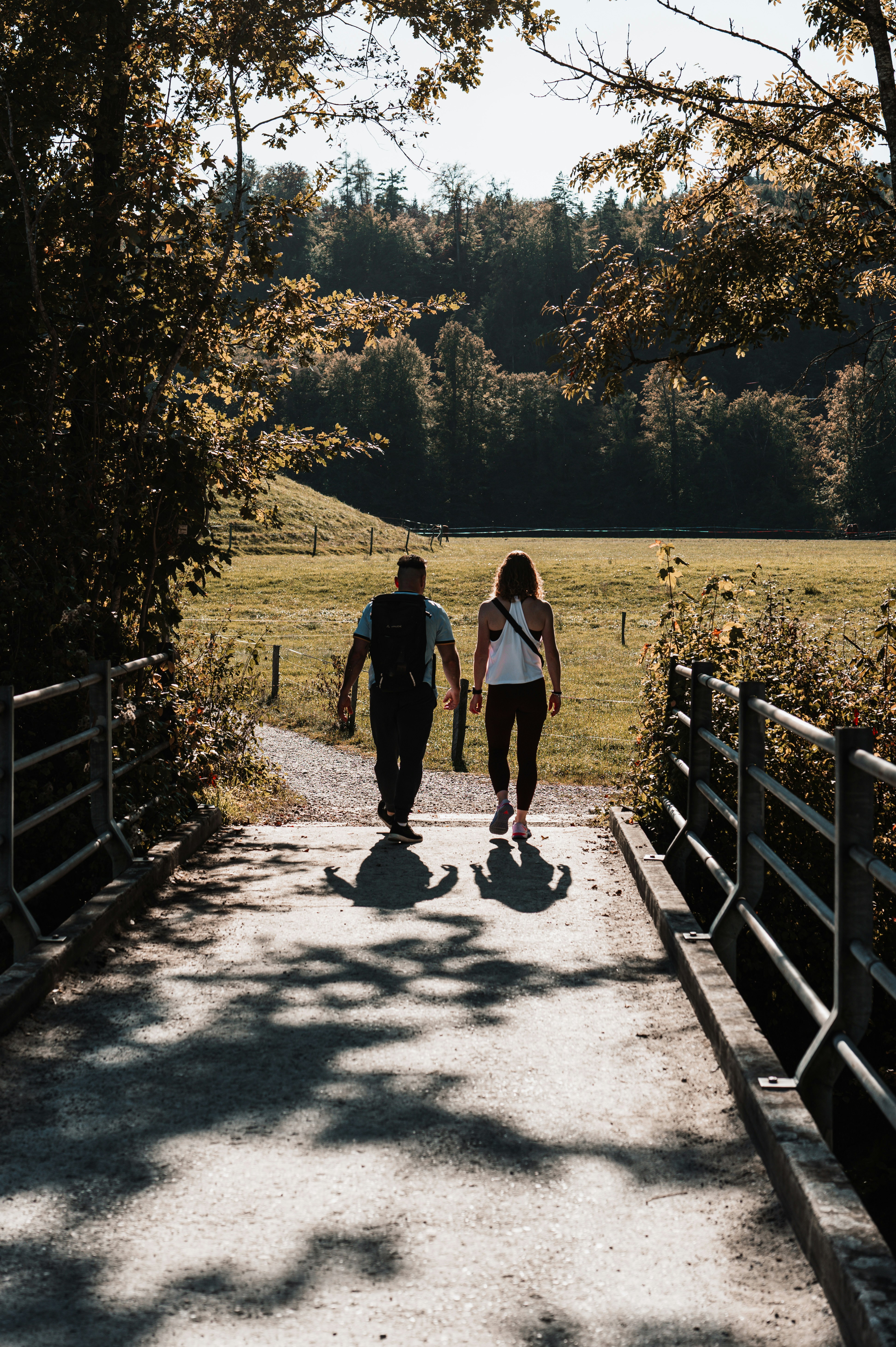 Two people walking across a concrete bridge toward a sunlit meadow