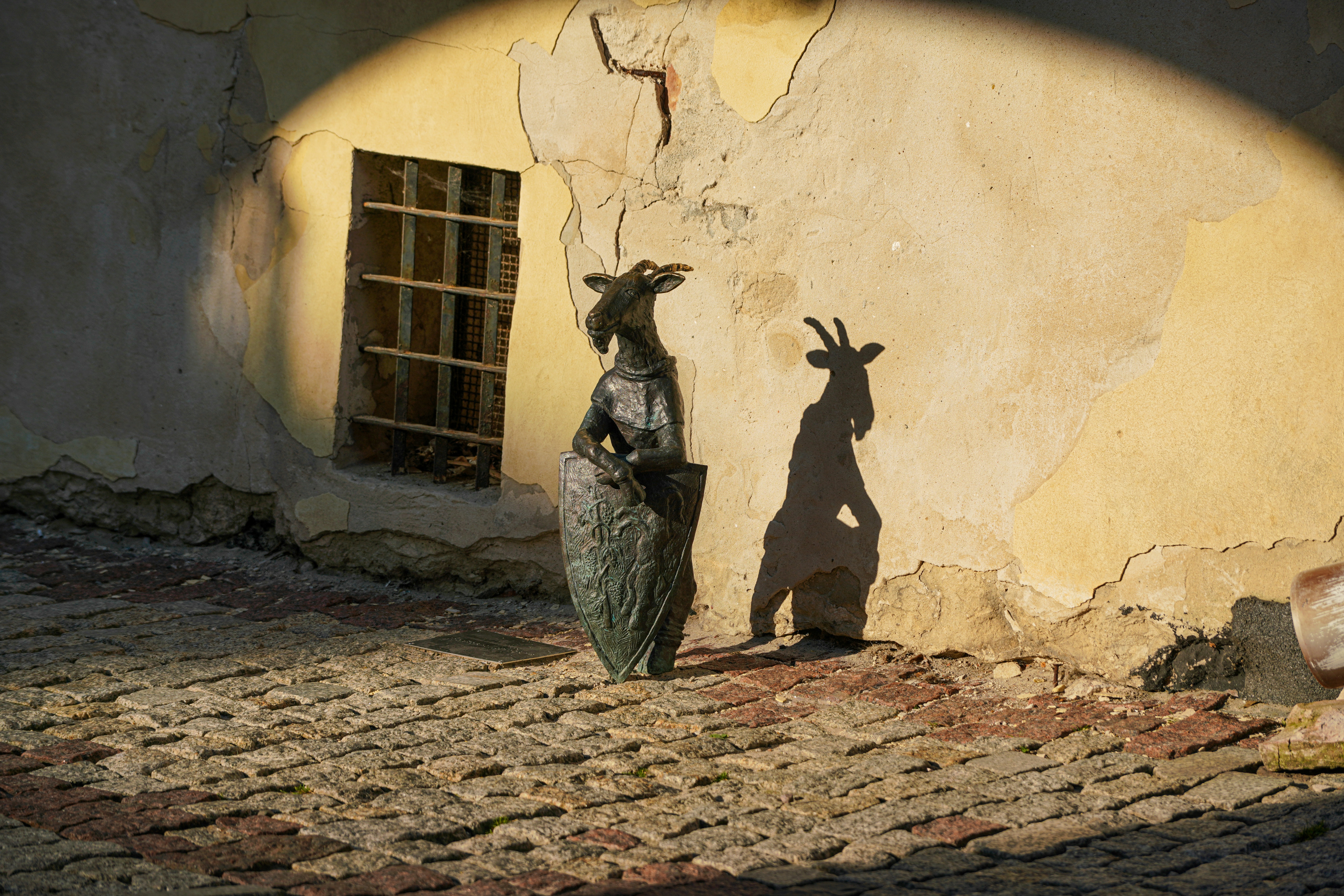A bronze goat statue holding a shield against a weathered yellow wall