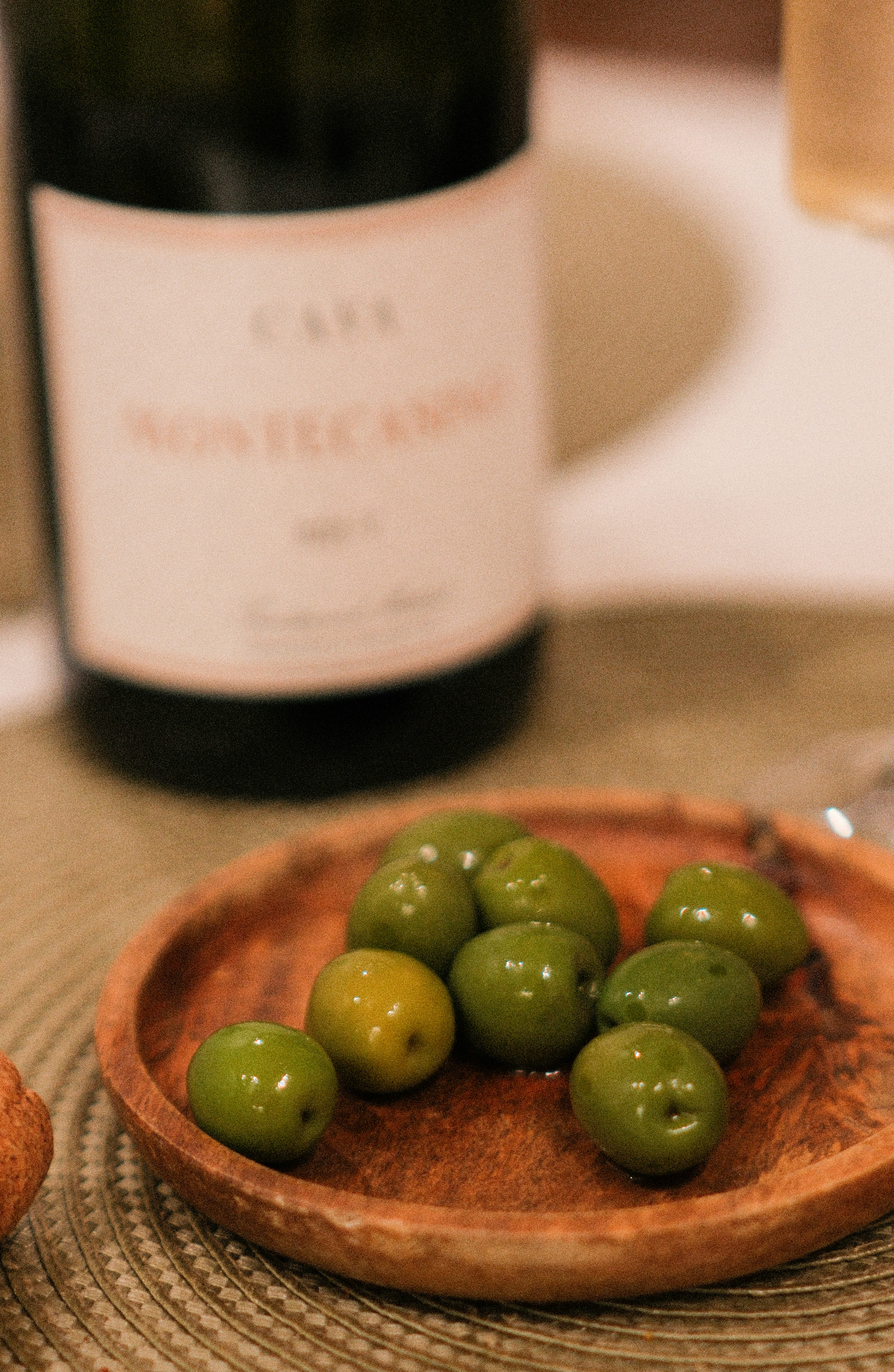 Green olives on a small wooden plate with a wine bottle in the background