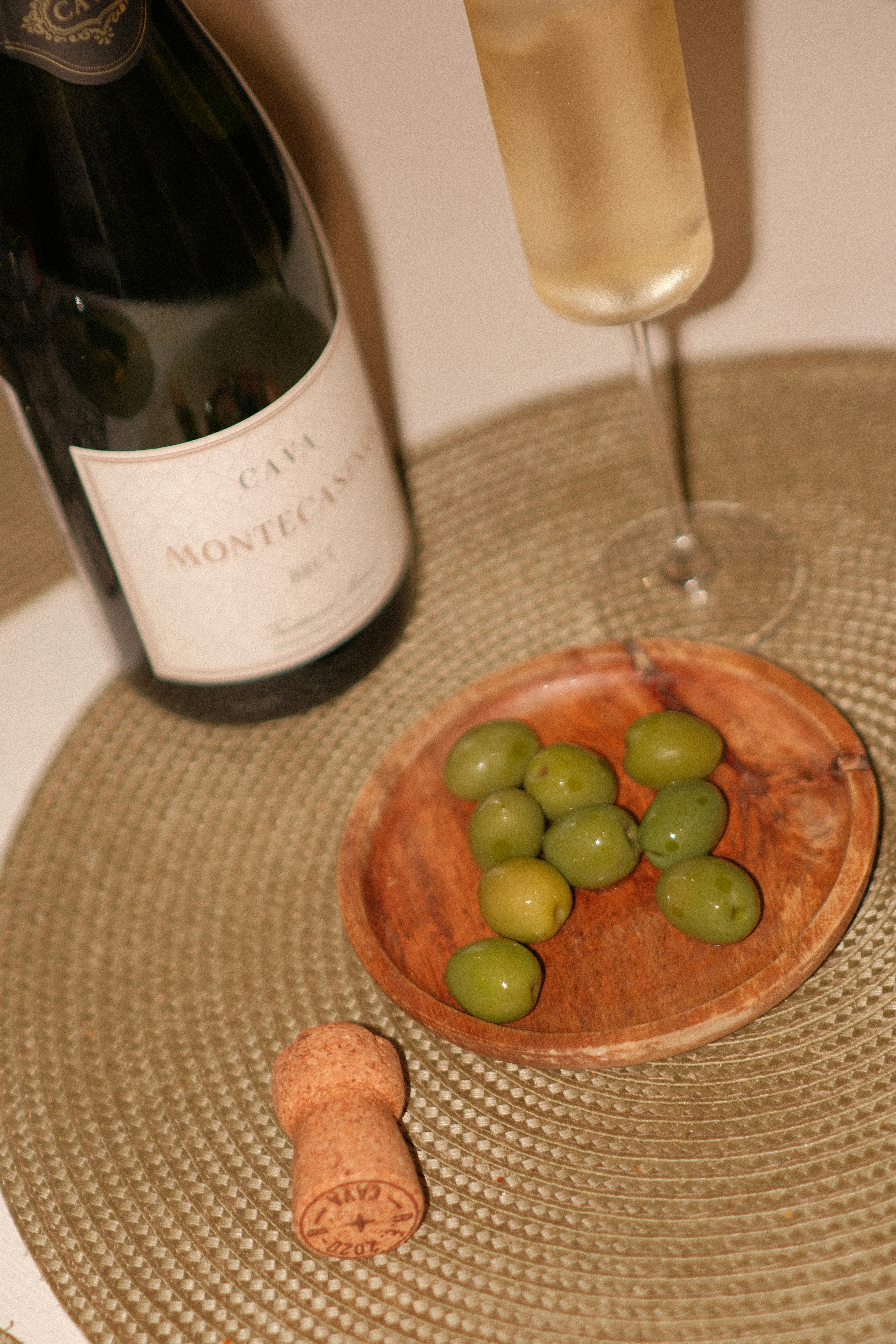 Green olives on a wooden plate beside a bottle of Cava and a glass