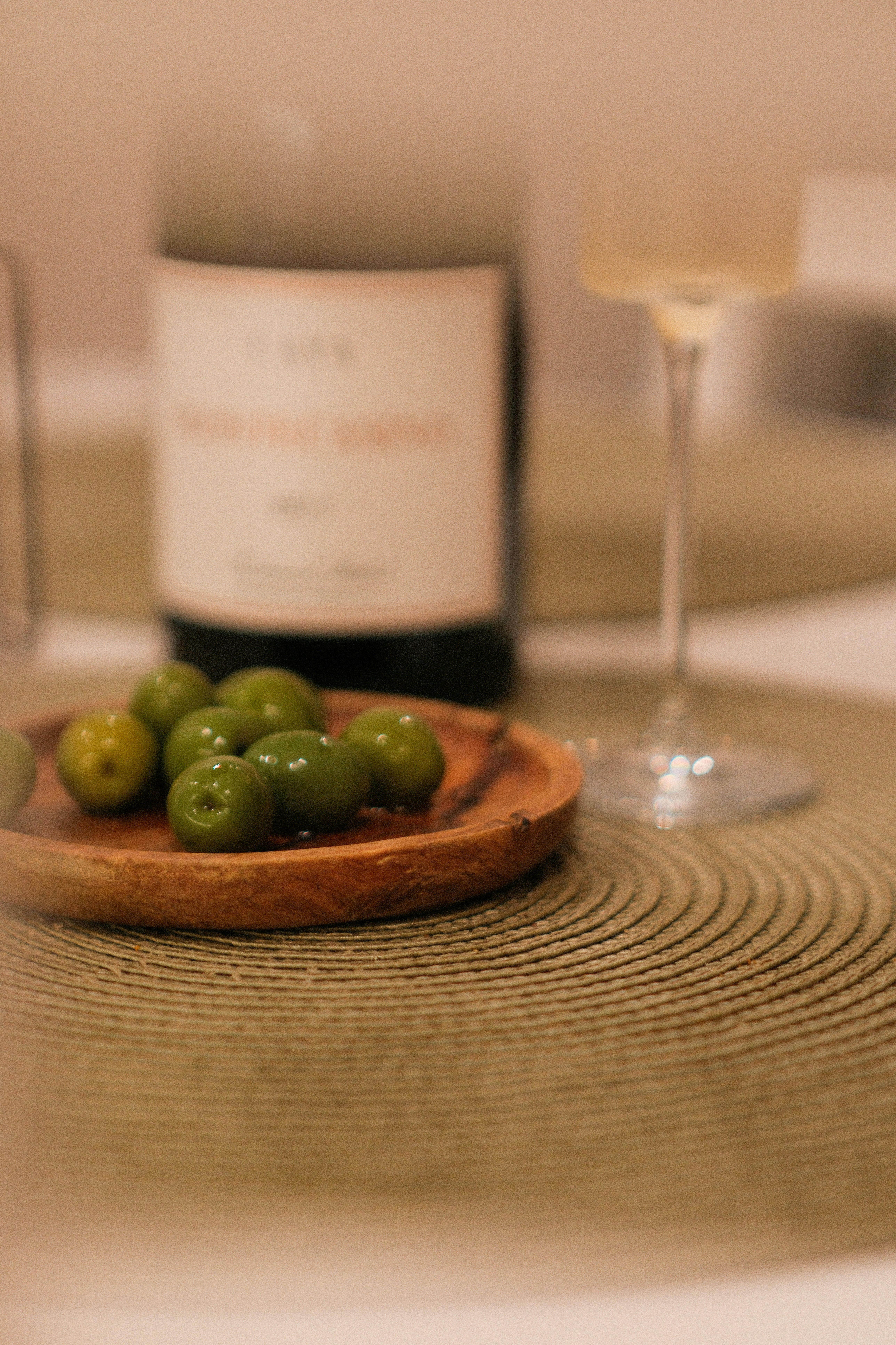 Green olives on a small wooden plate beside a wine bottle and glass