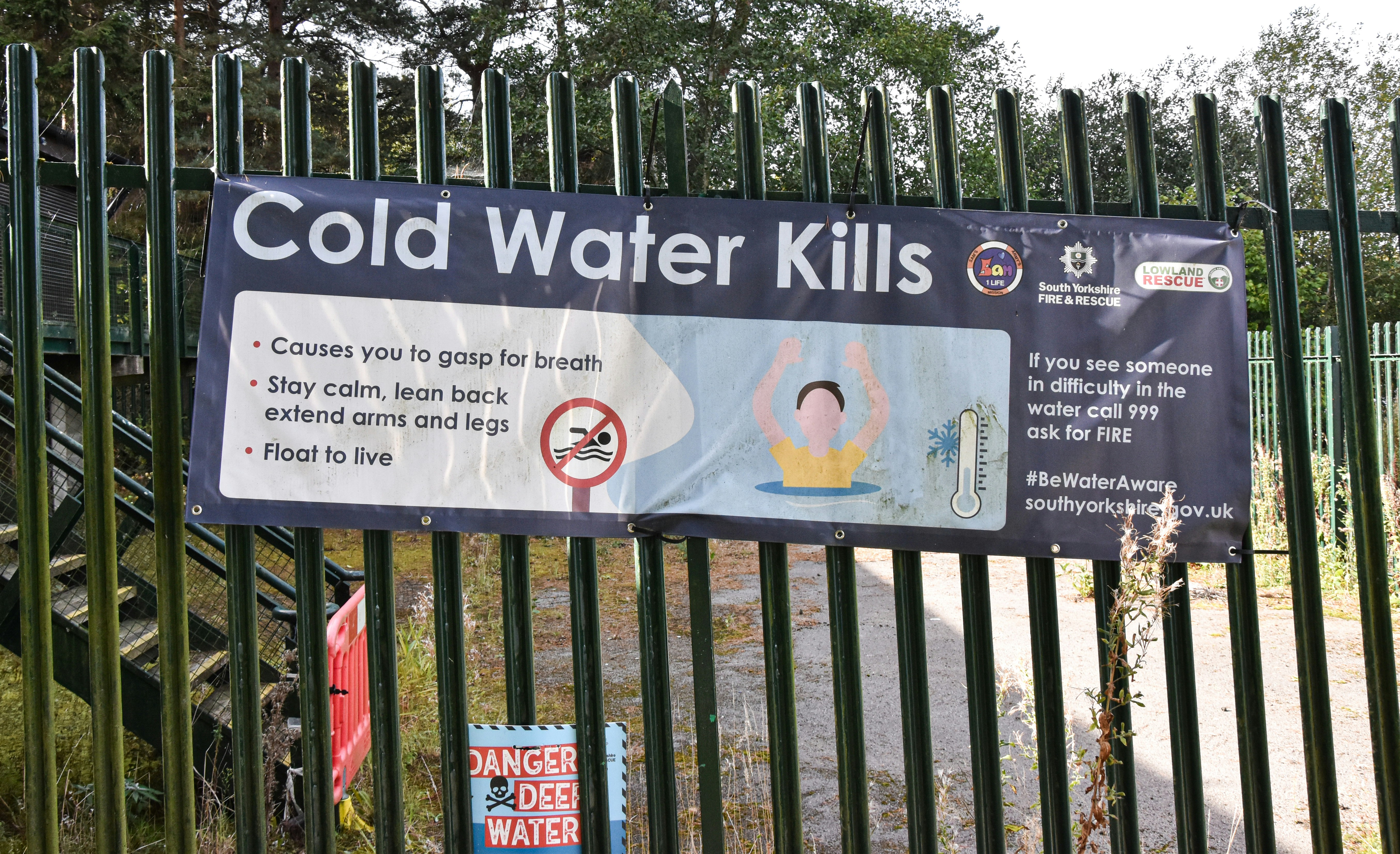 A safety banner reading Cold Water Kills attached to a green metal fence