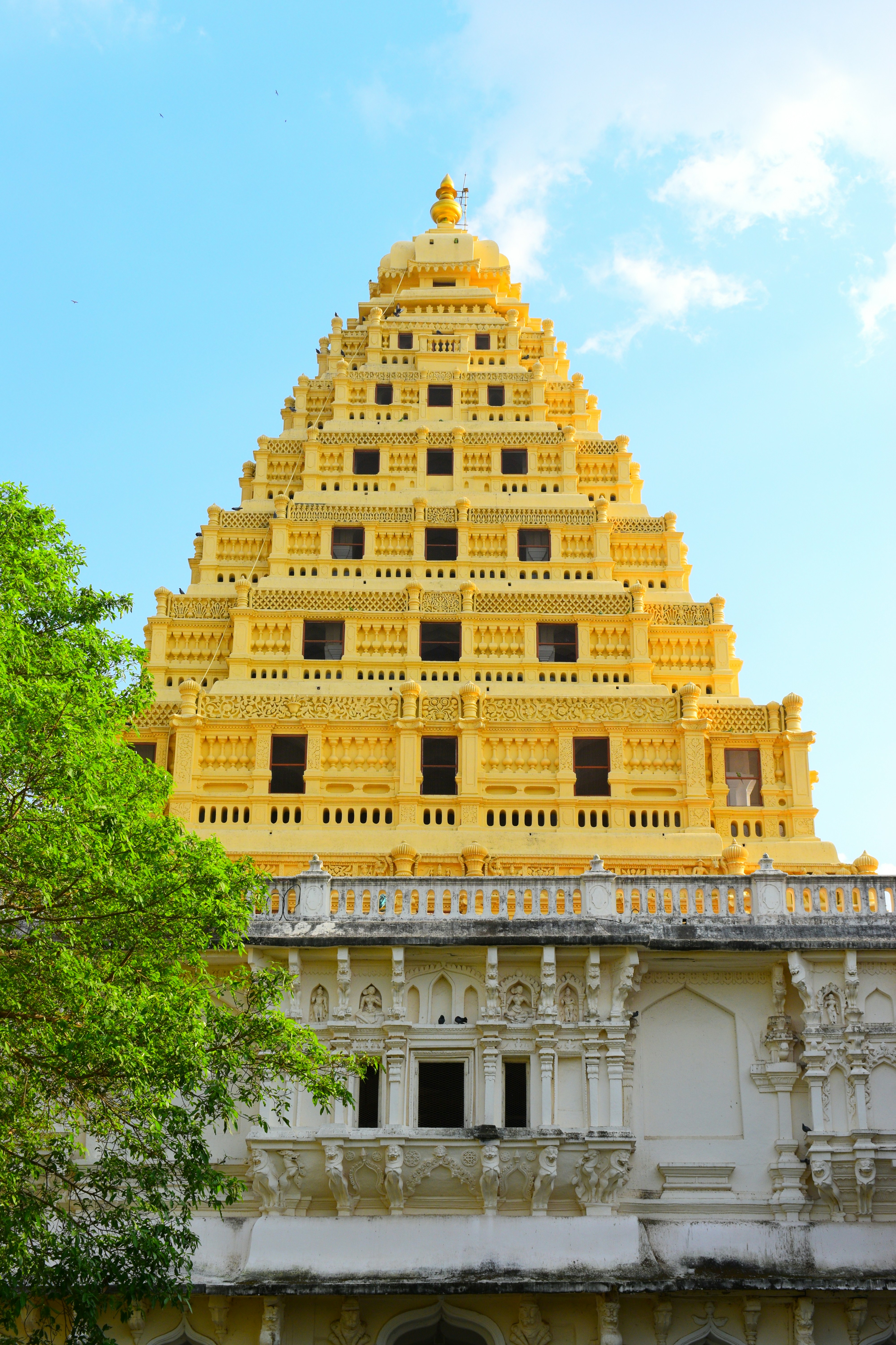 A tiered yellow temple tower rising above a white stone base with green foliage