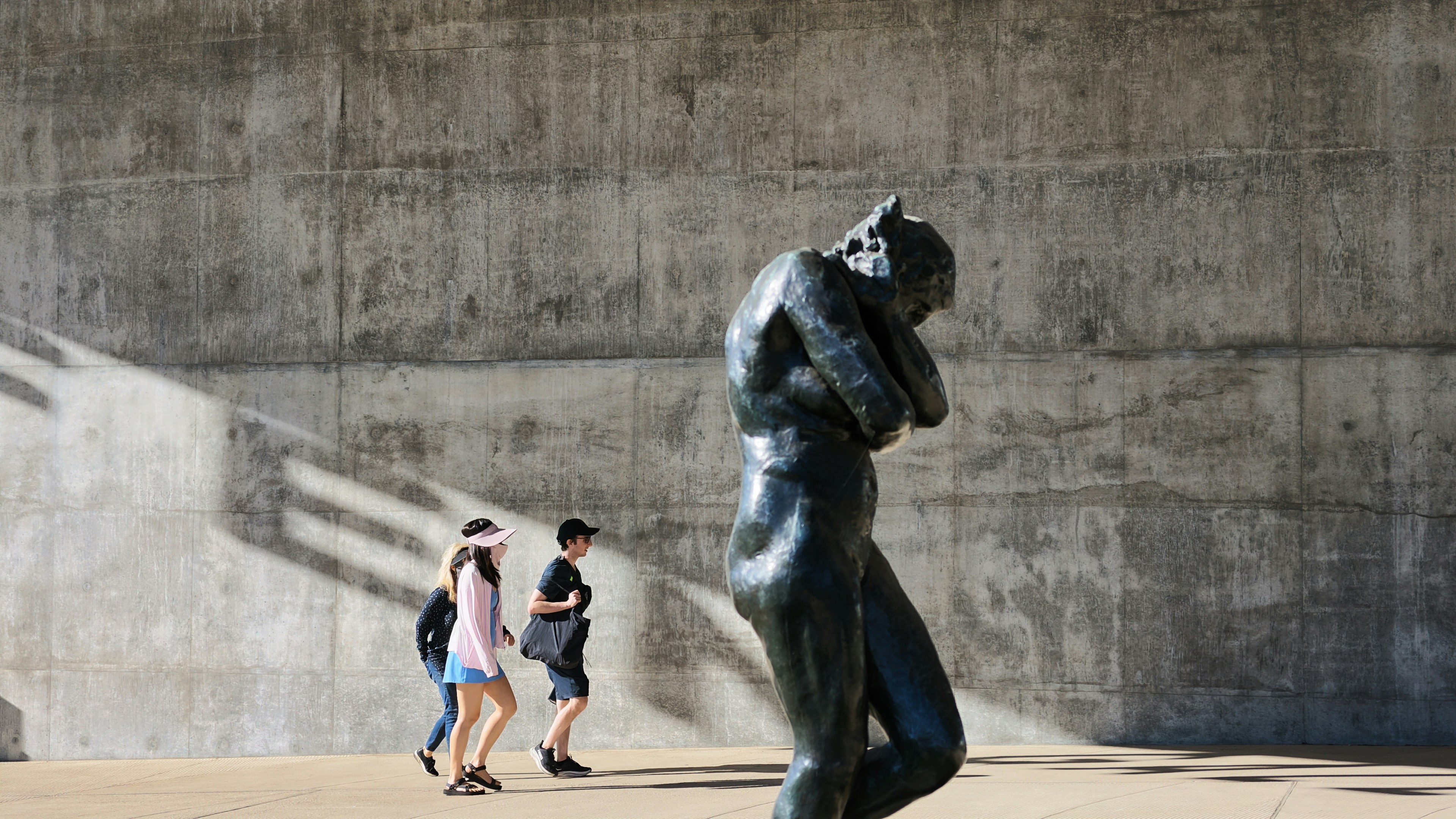 Three people walking past a bronze sculpture against a large concrete wall