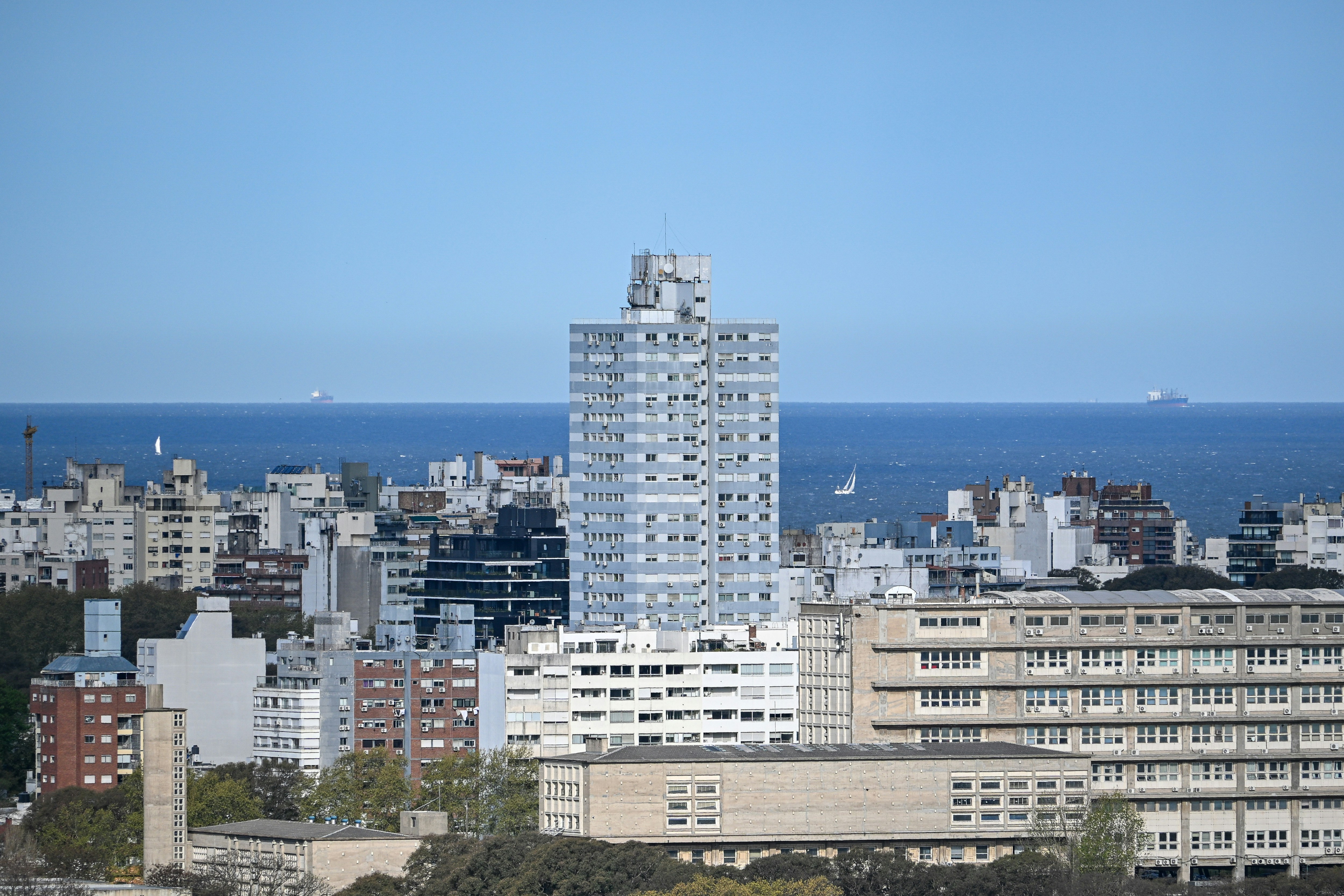A tall blue apartment building rising above a dense city skyline near the ocean