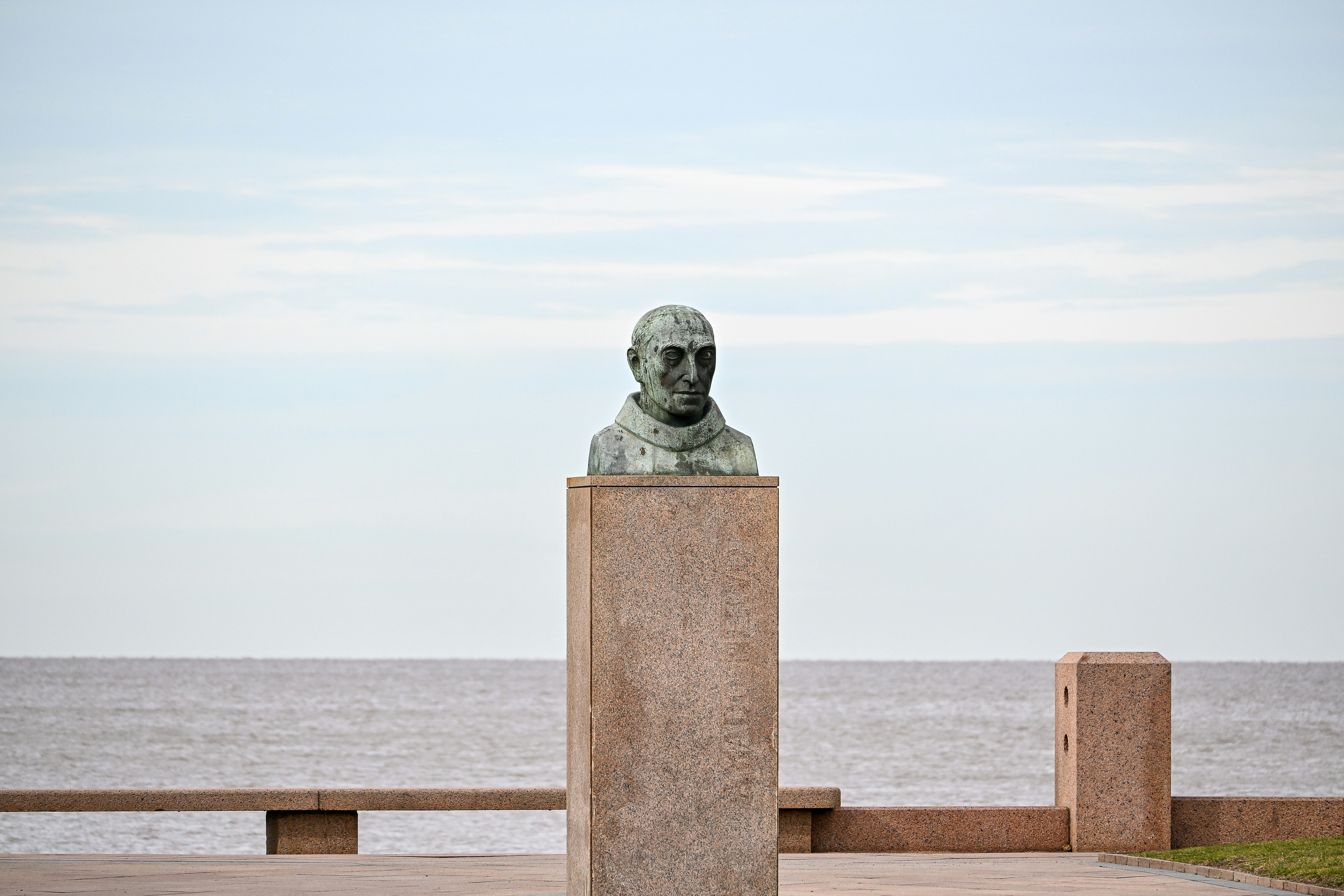 A bronze bust of a man on a tall stone pedestal by the sea