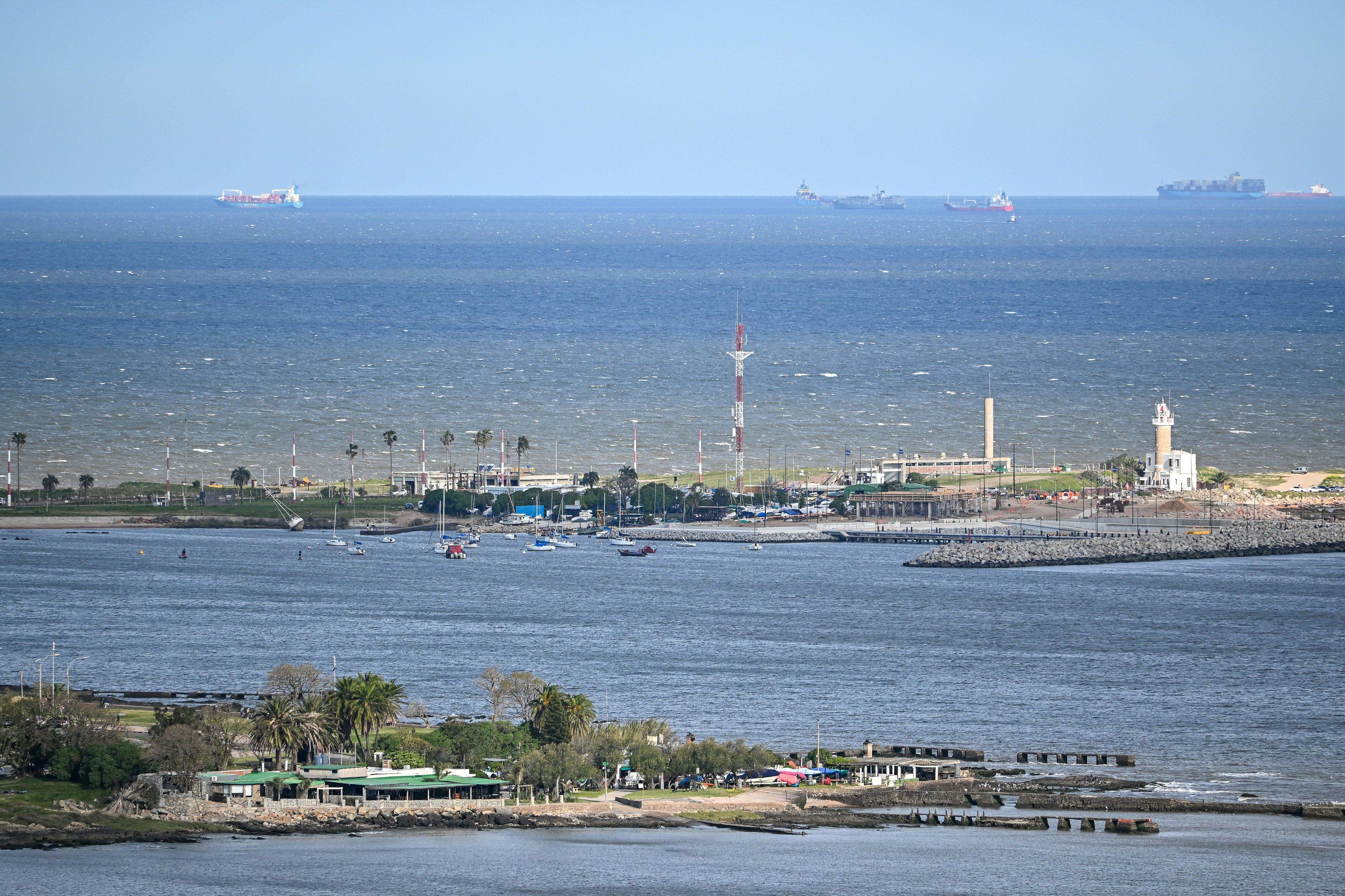 A lighthouse and marina on a rocky coastline with ships on the horizon