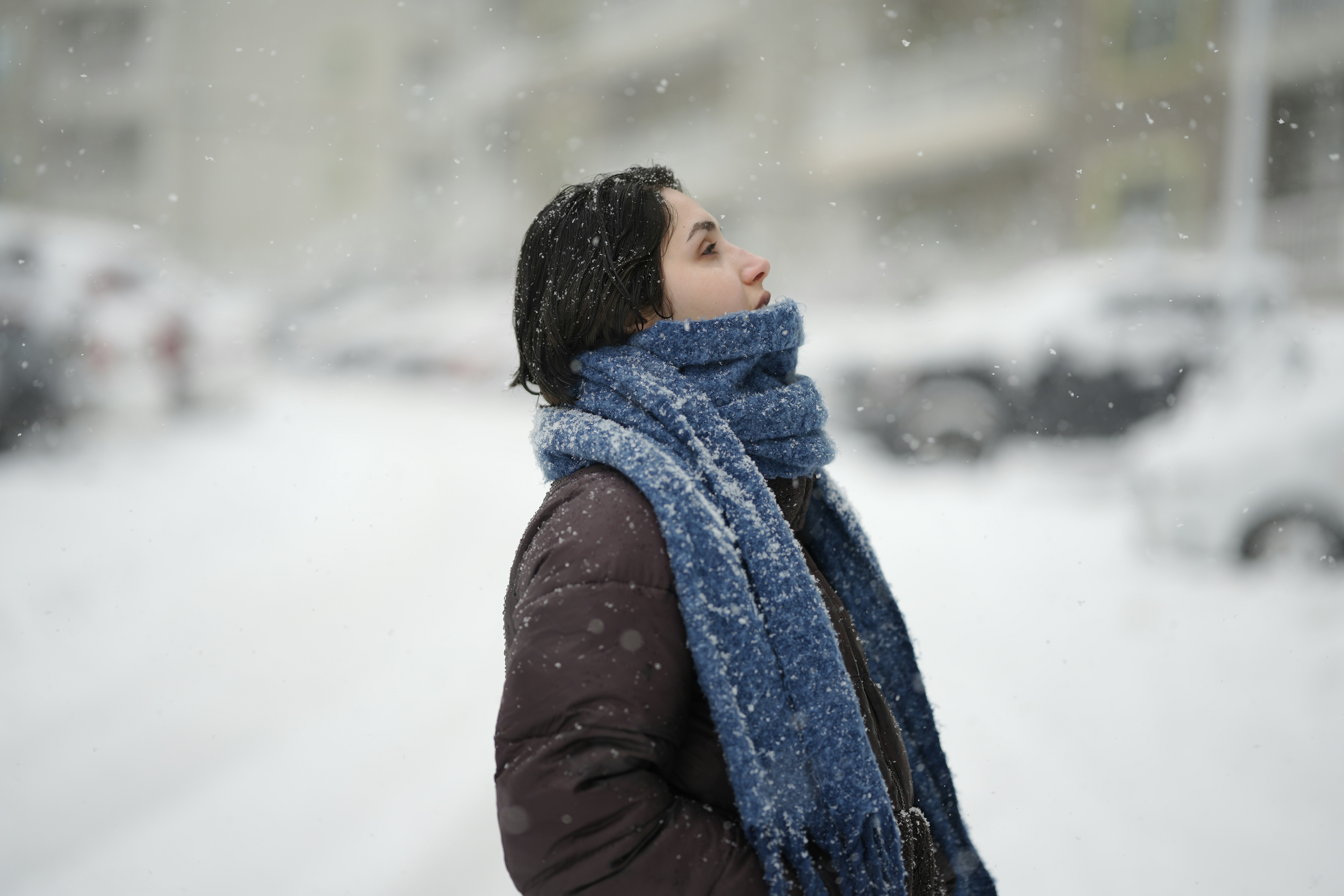 雪の中、暗いジャケットと青いマフラーを身に着けた女性が見上げている