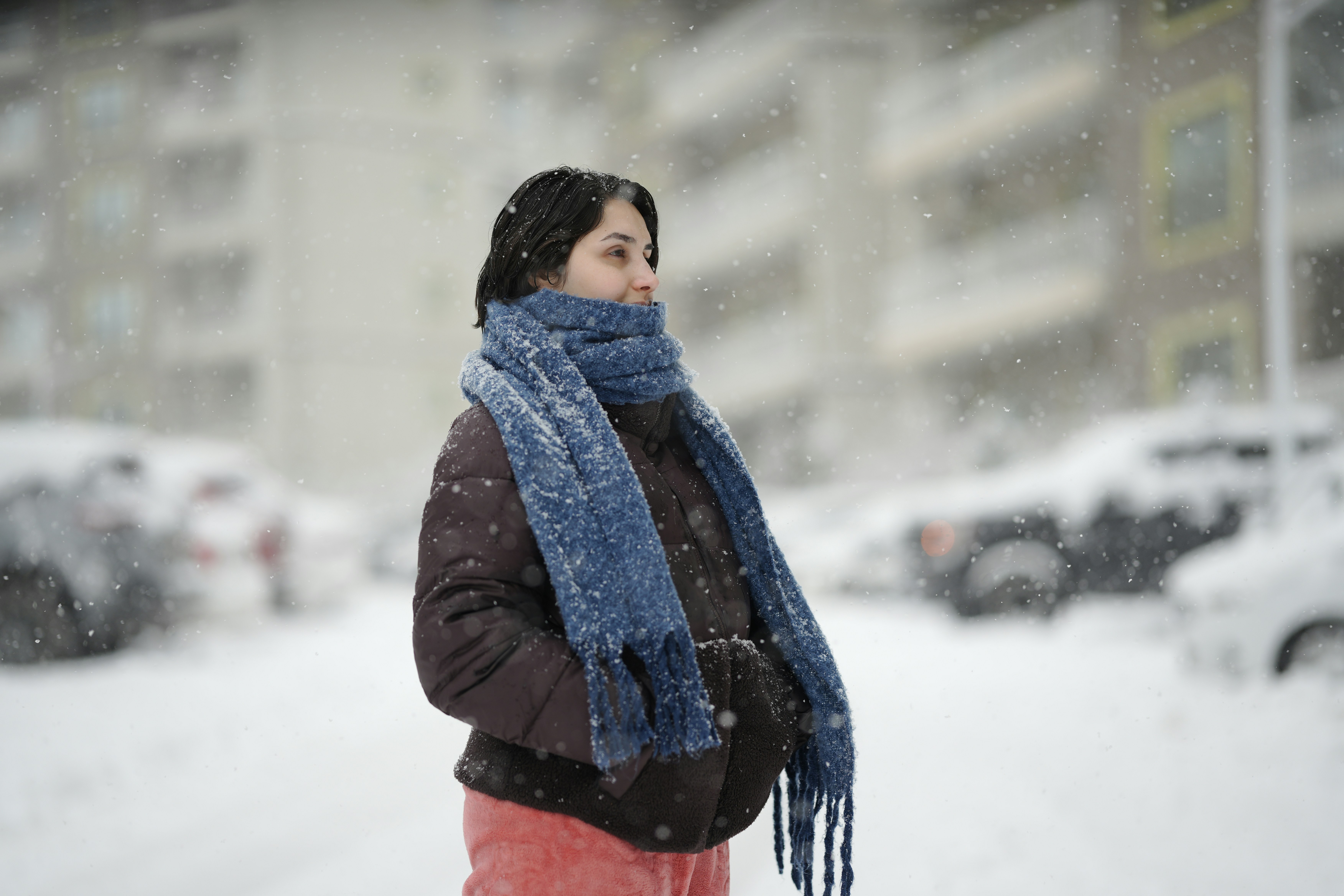雪の降る中、茶色のパファージャケットと青いマフラーを身に着けた女性