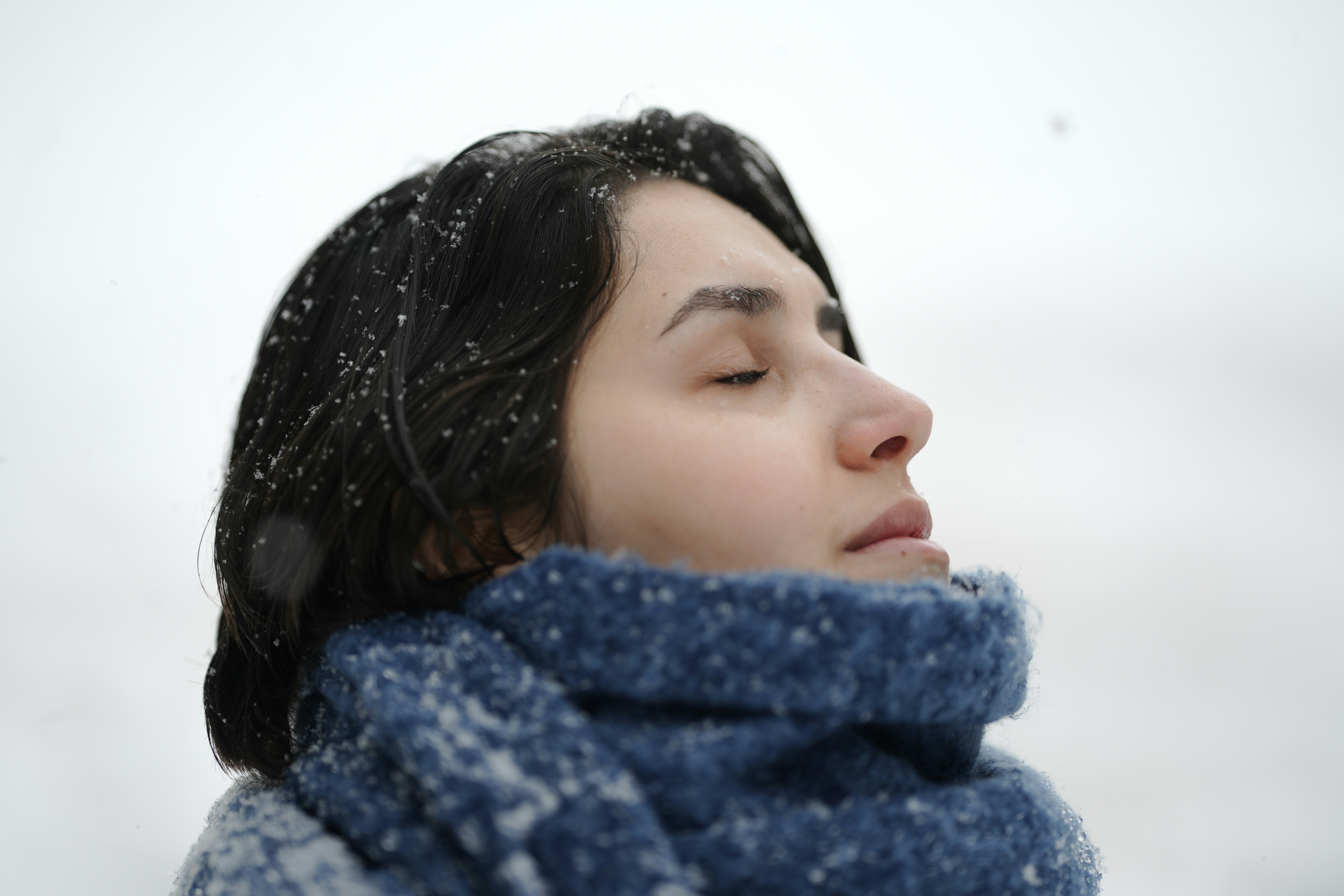 Uma mulher de cabelos escuros e olhos fechados na neve caindo, usando um cachecol azul