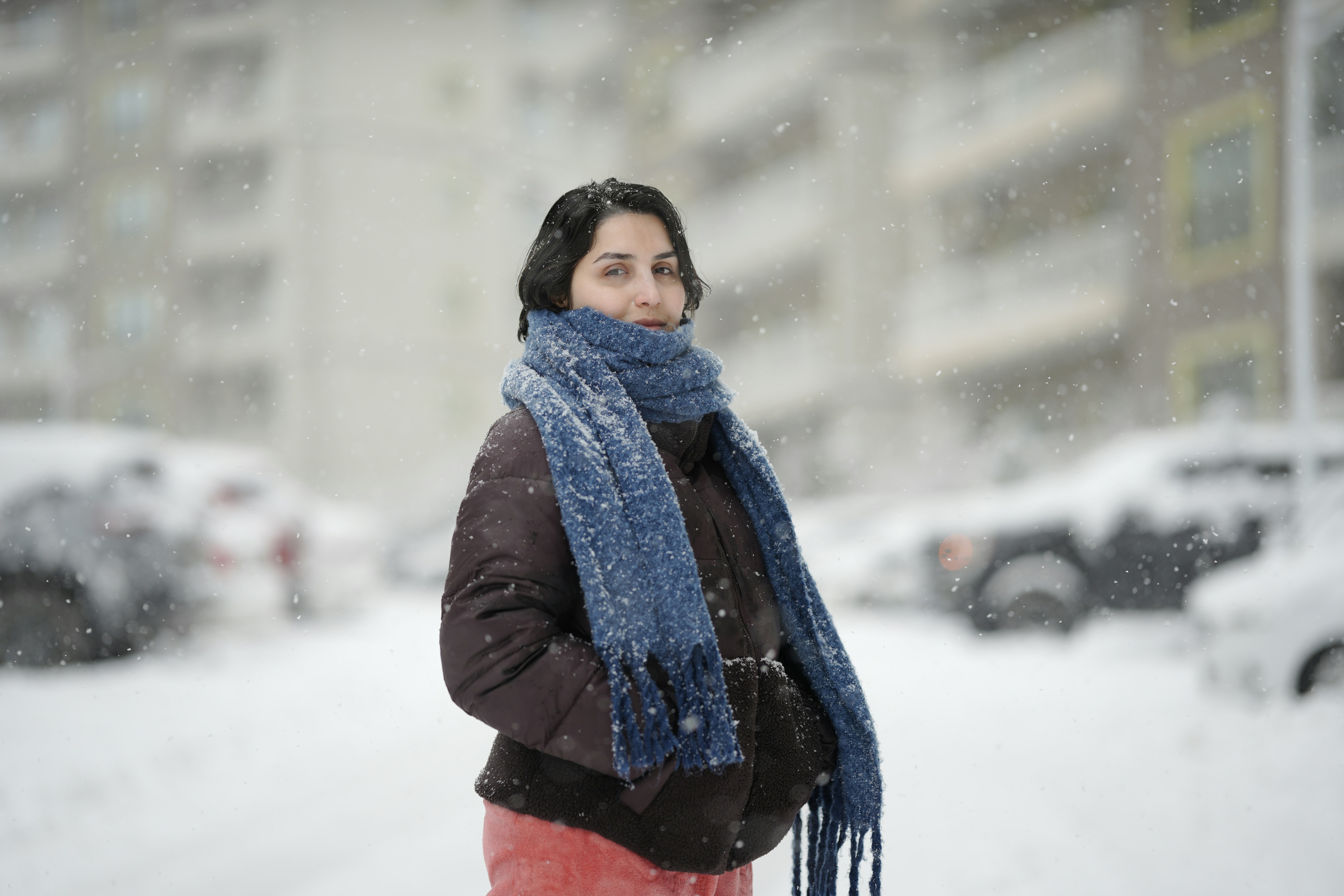 Uma mulher de jaqueta marrom e cachecol azul parada na neve caindo