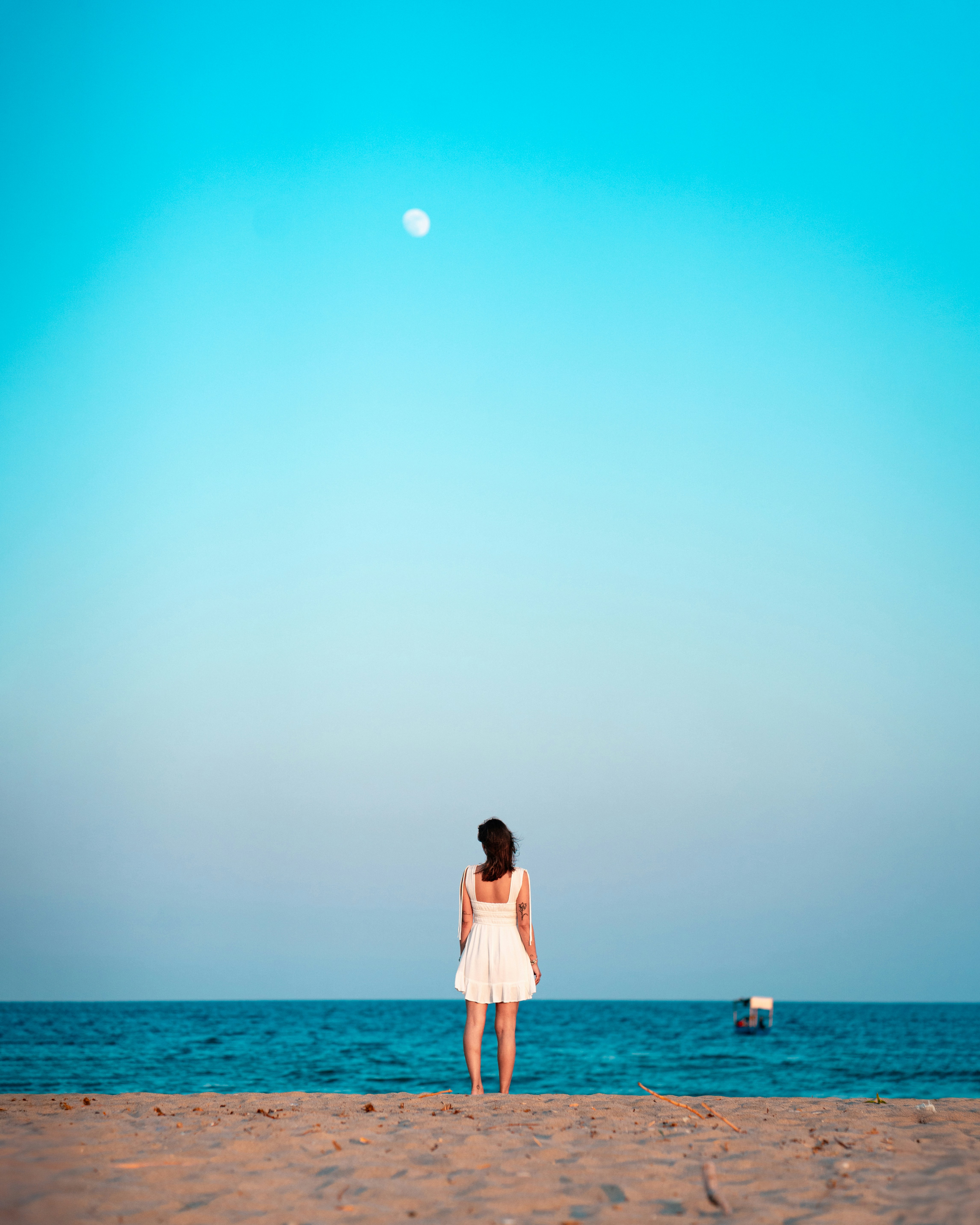 A woman in a white dress on a sandy beach facing the ocean