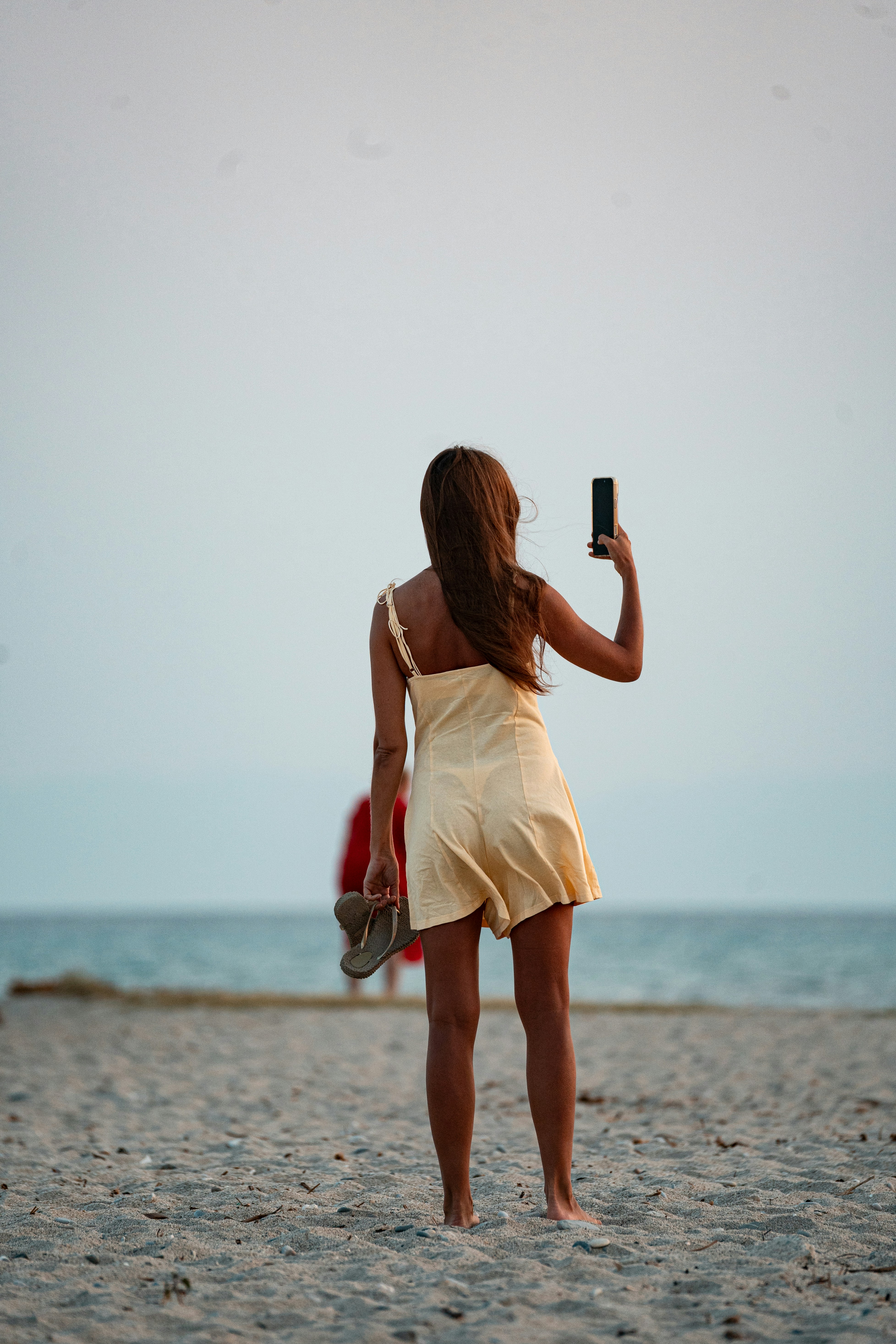 A woman in a yellow dress holding a phone and sandals on a beach