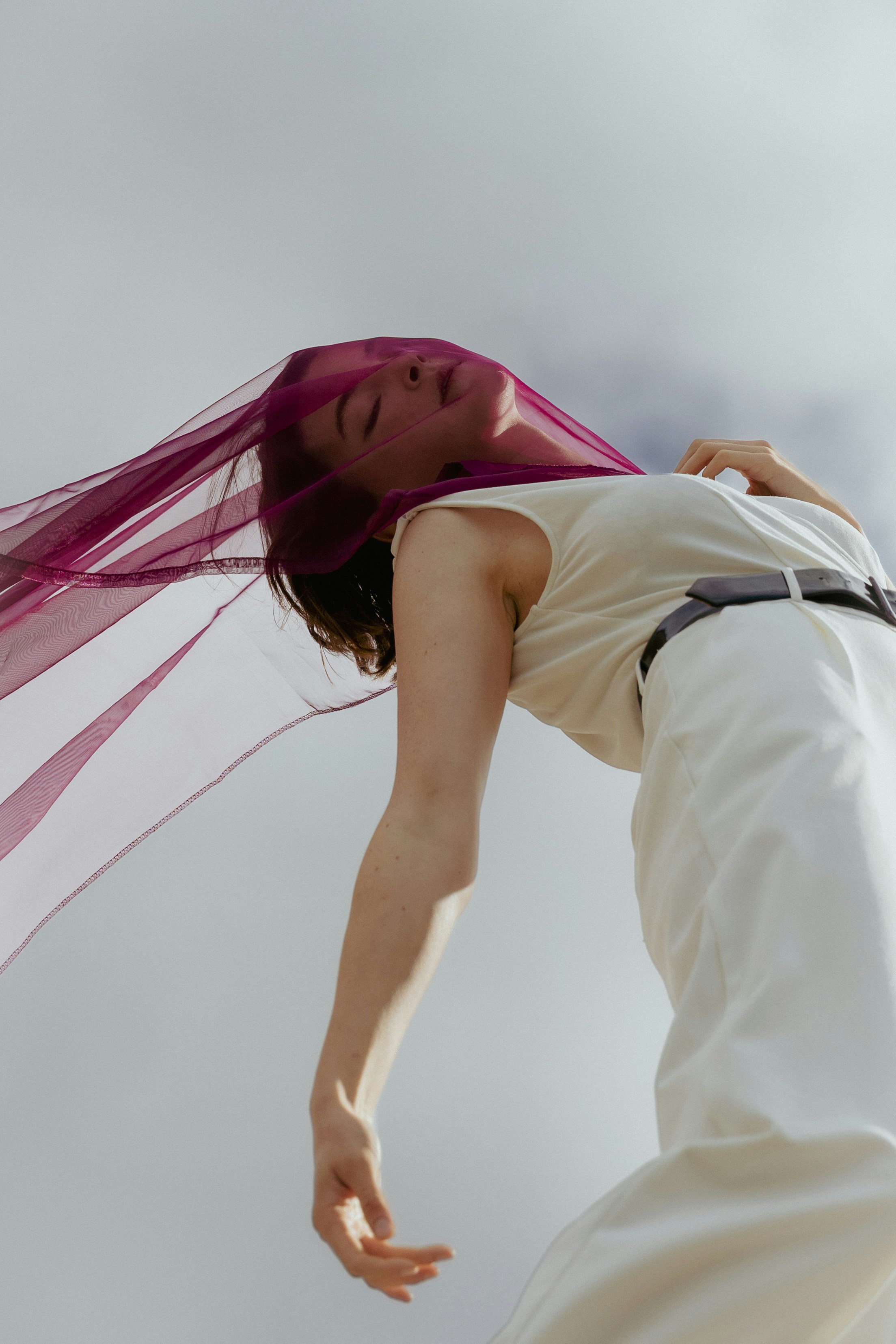 A person in a white jumpsuit wearing a sheer purple veil against a sky