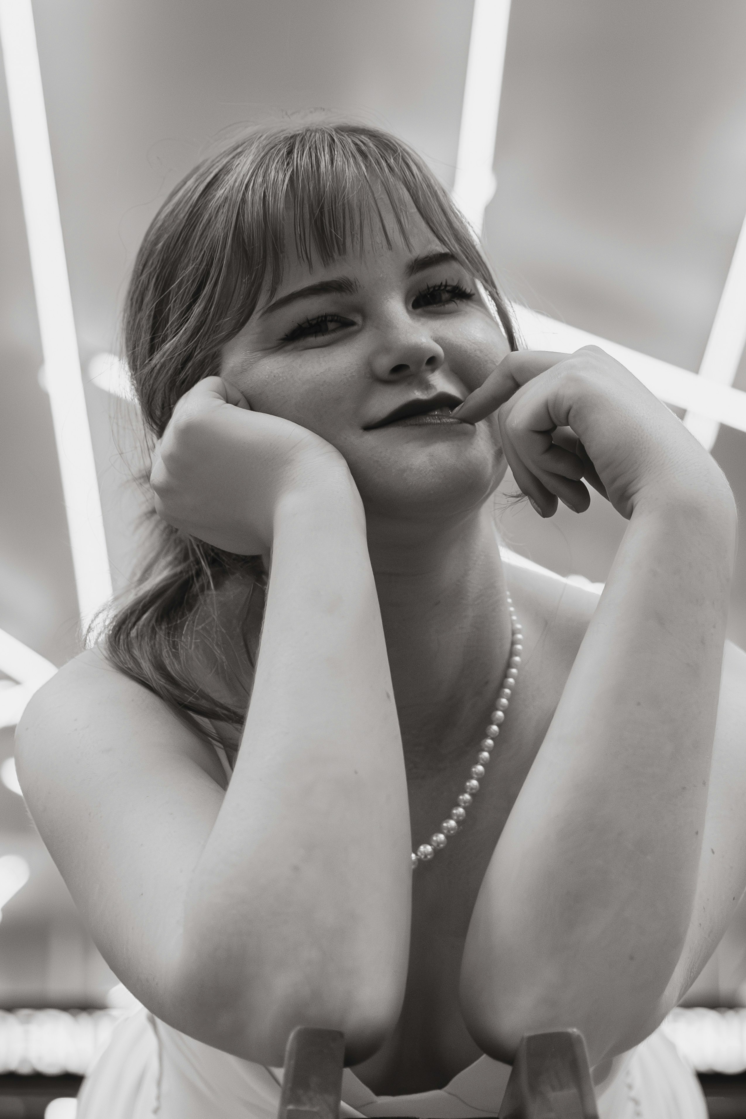 Une femme à la frange portant un collier de perles, les mains posées sur son menton