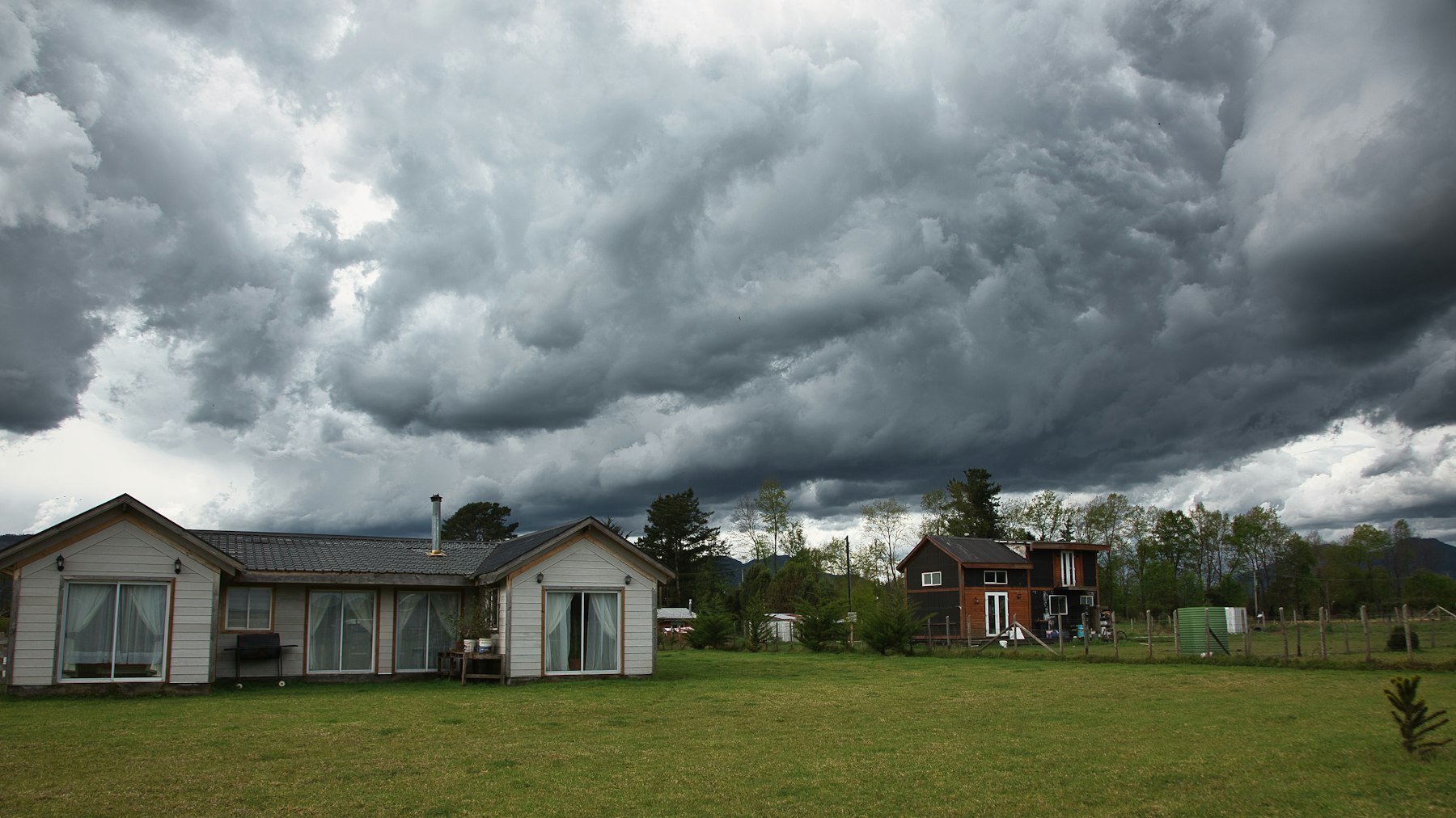Two farmhouses on open grassland beneath towering dark storm clouds
