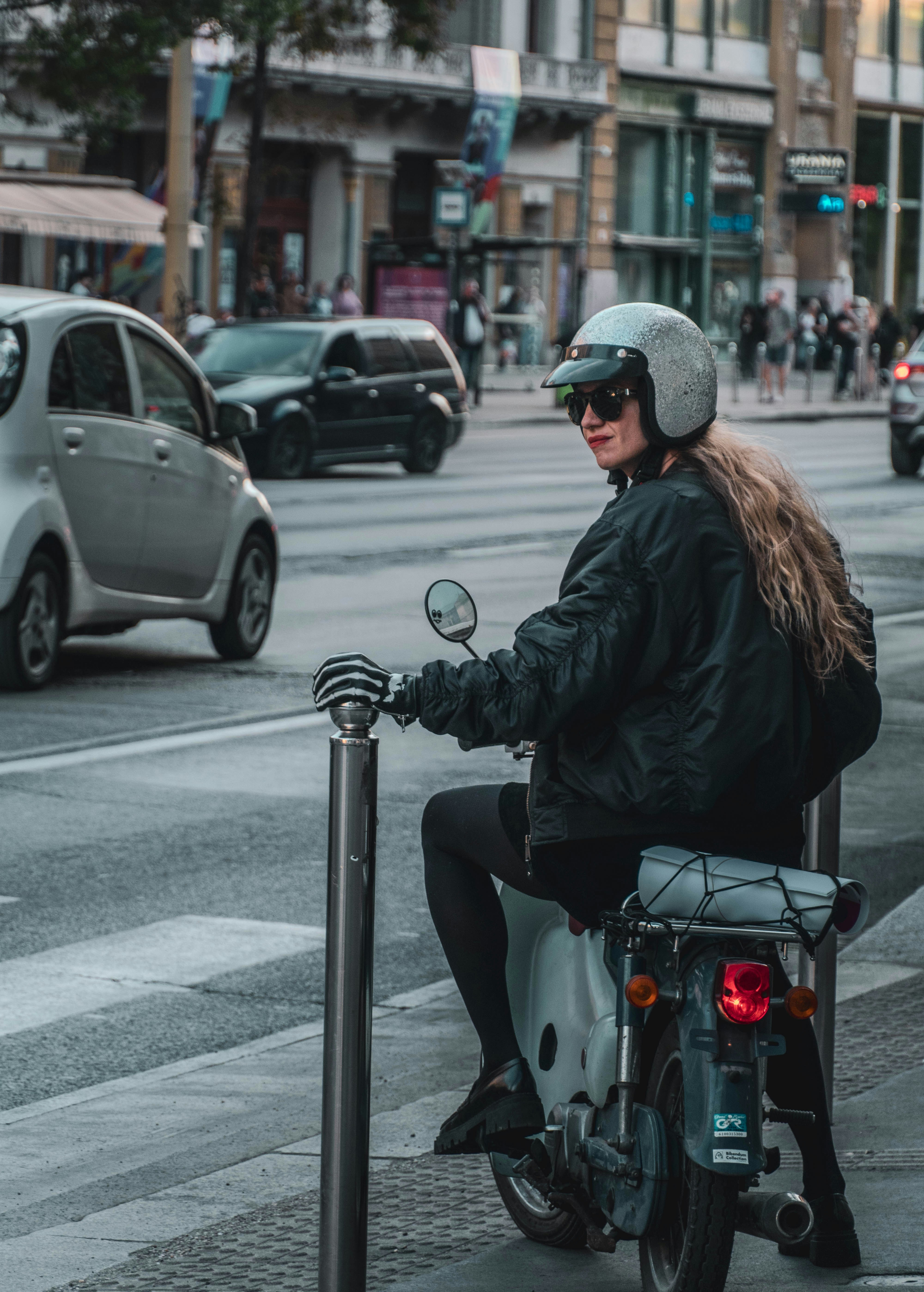 A woman wearing a silver helmet on a moped beside a city street