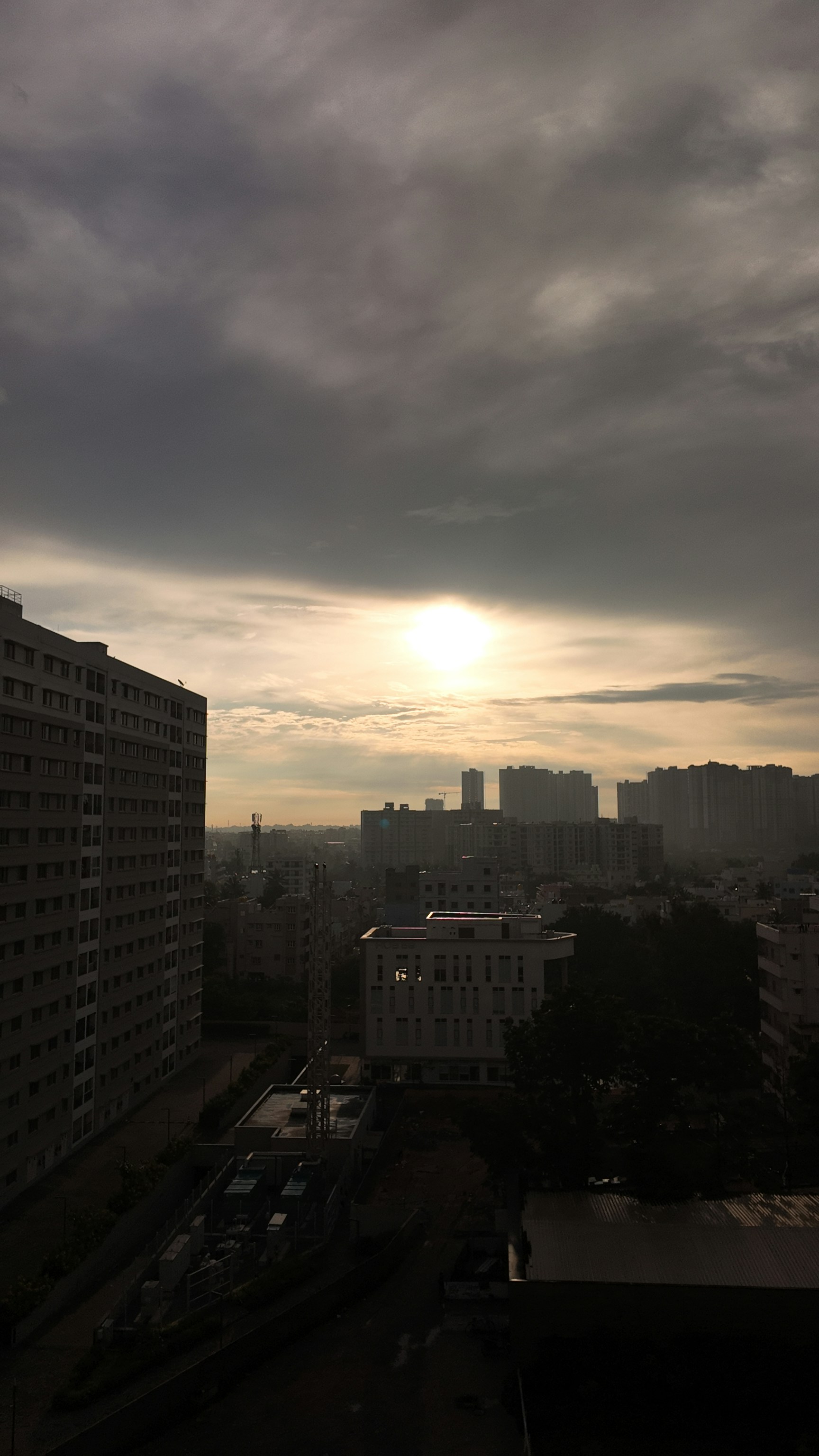 Dusk light over a clustered urban skyline with a bright sun low on the horizon; foreground shows mid-rise buildings and a quiet street, creating a moody, atmospheric city scene. This is a photographic capture of a real urban landscape.