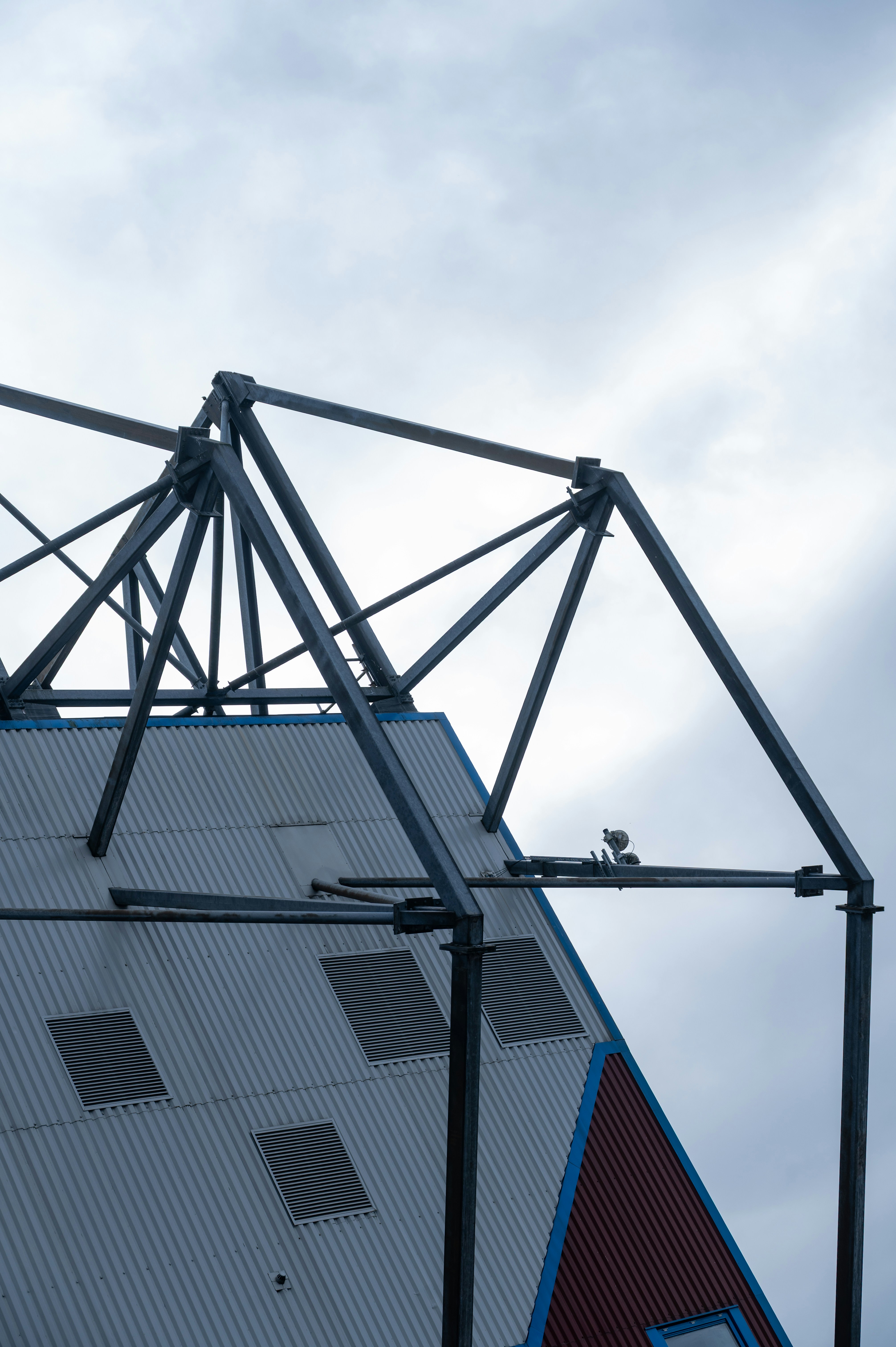 A steel truss framework above a corrugated metal roof against a cloudy sky