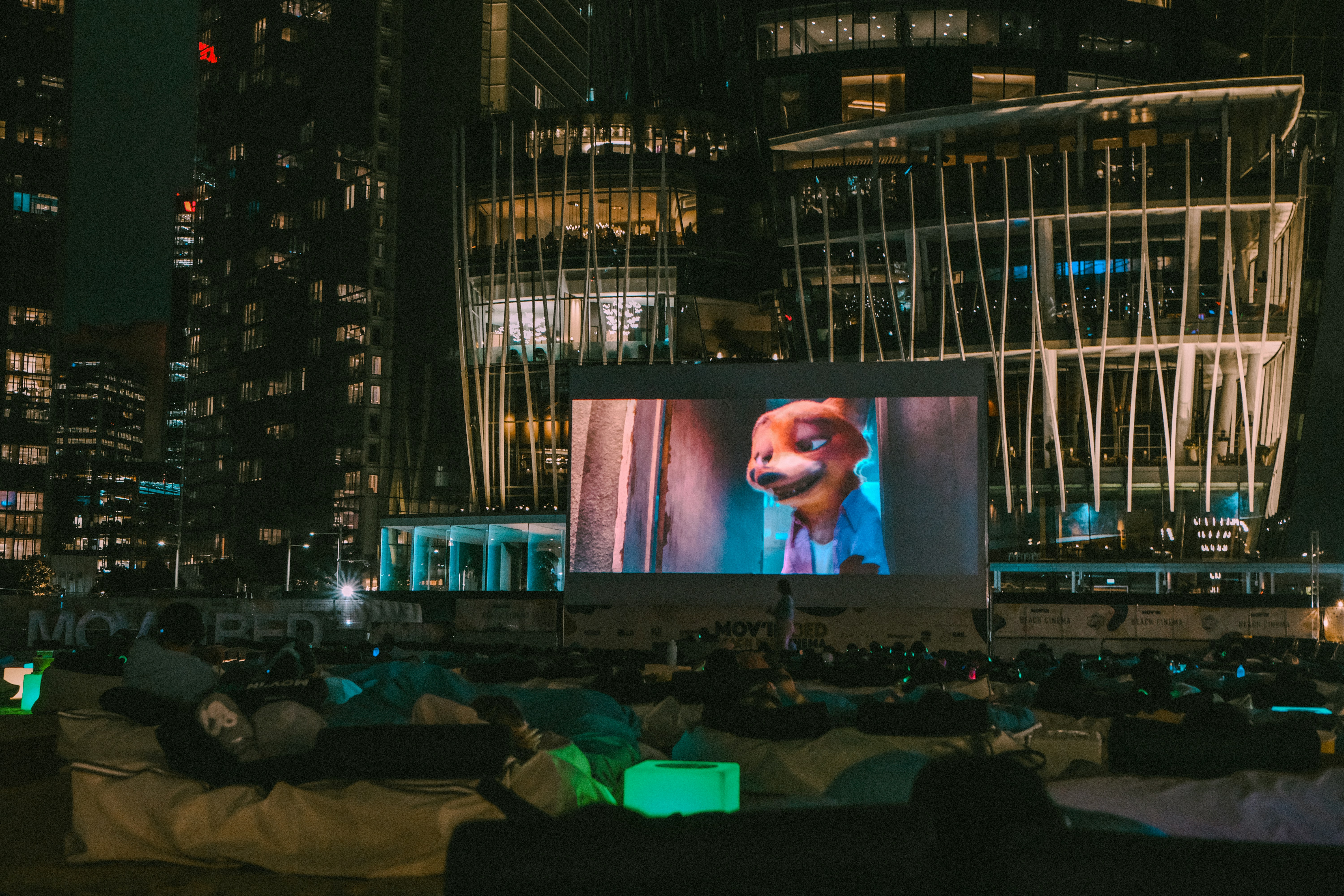 Des spectateurs allongés sur des poufs regardent un film d’animation projeté sur un écran géant en plein air, de nuit, au pied de tours