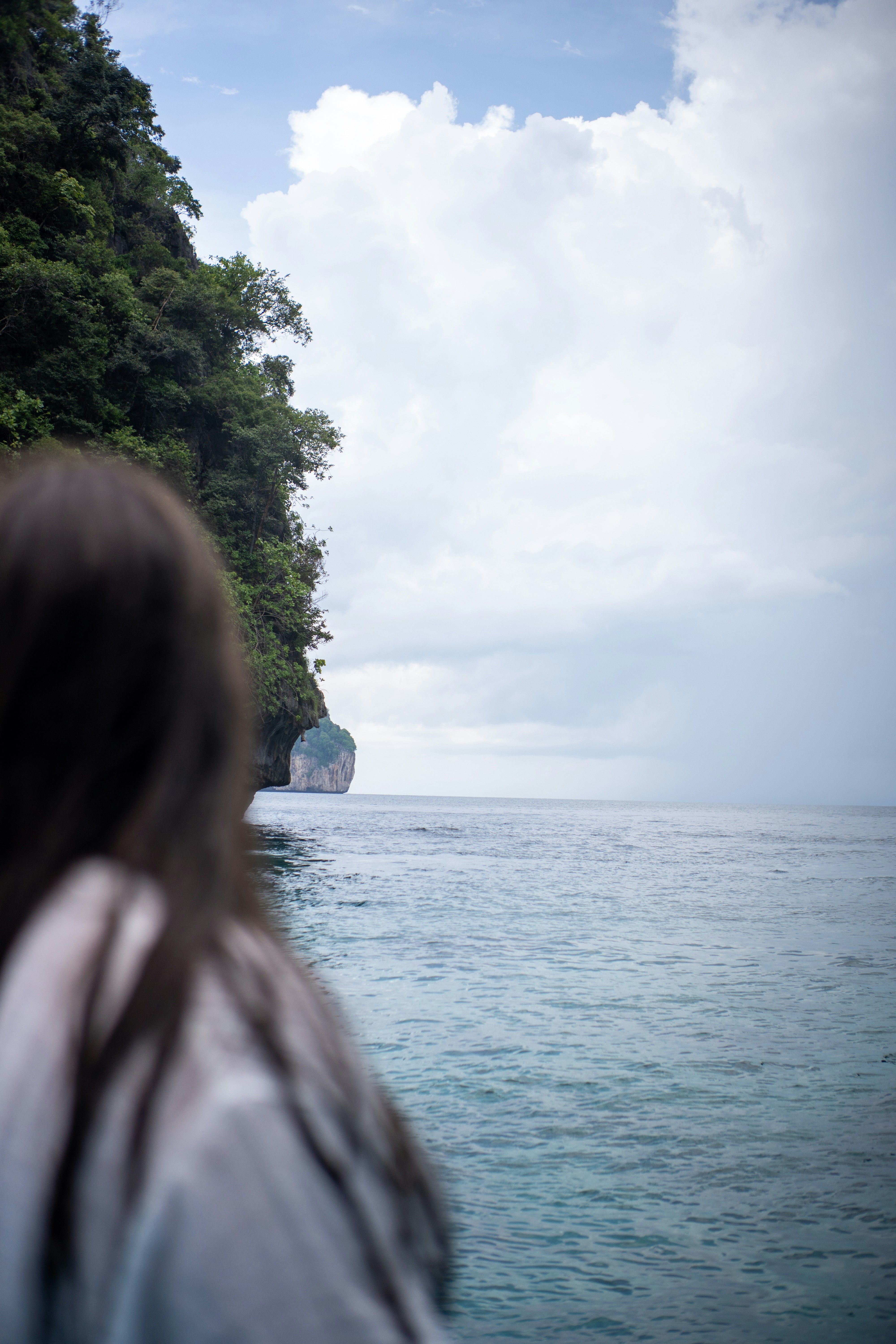 A person looking toward a limestone cliff rising from the sea under cloudy skies