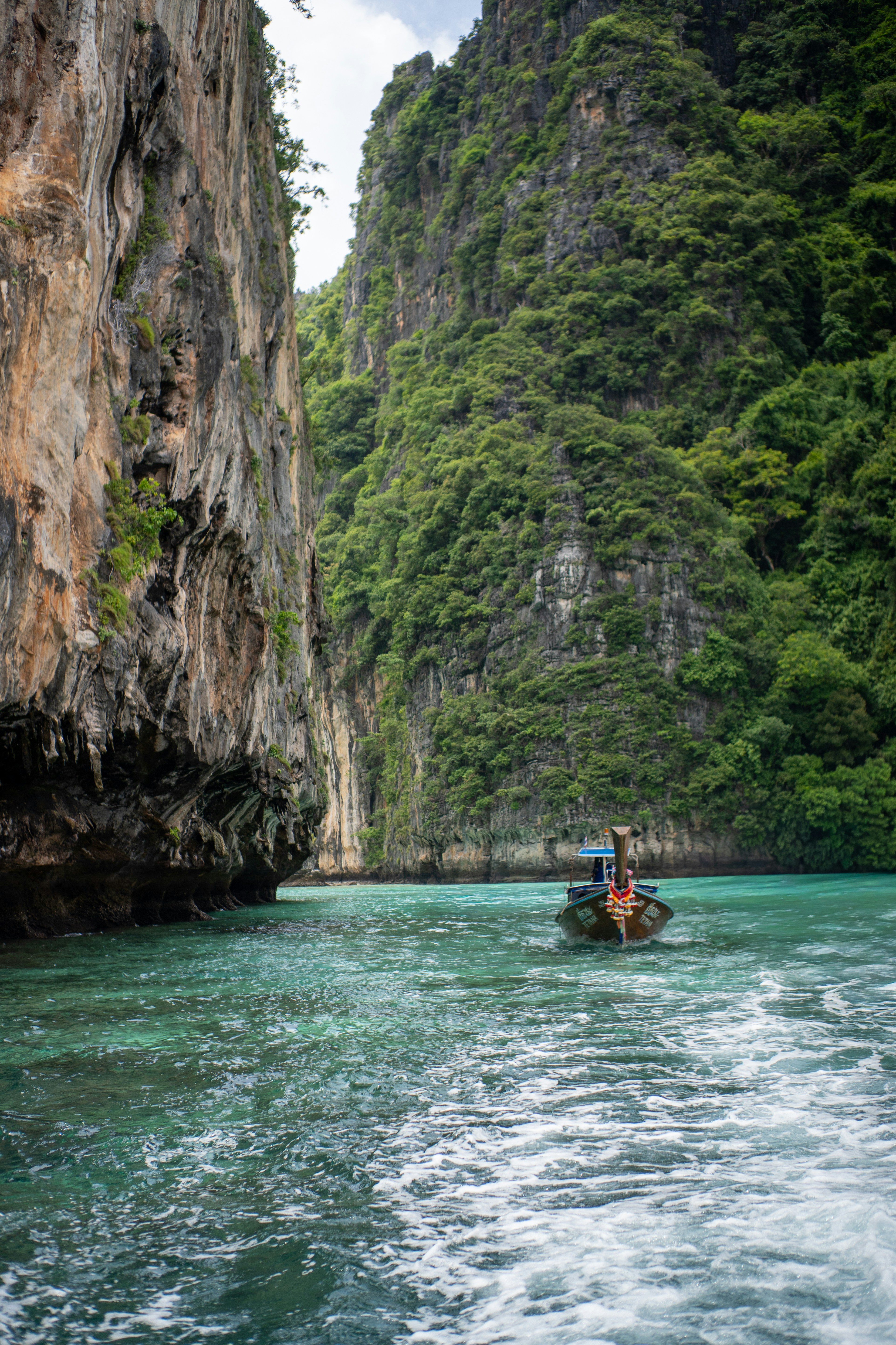 A long-tail boat moving through turquoise water between towering limestone cliffs in Thailand
