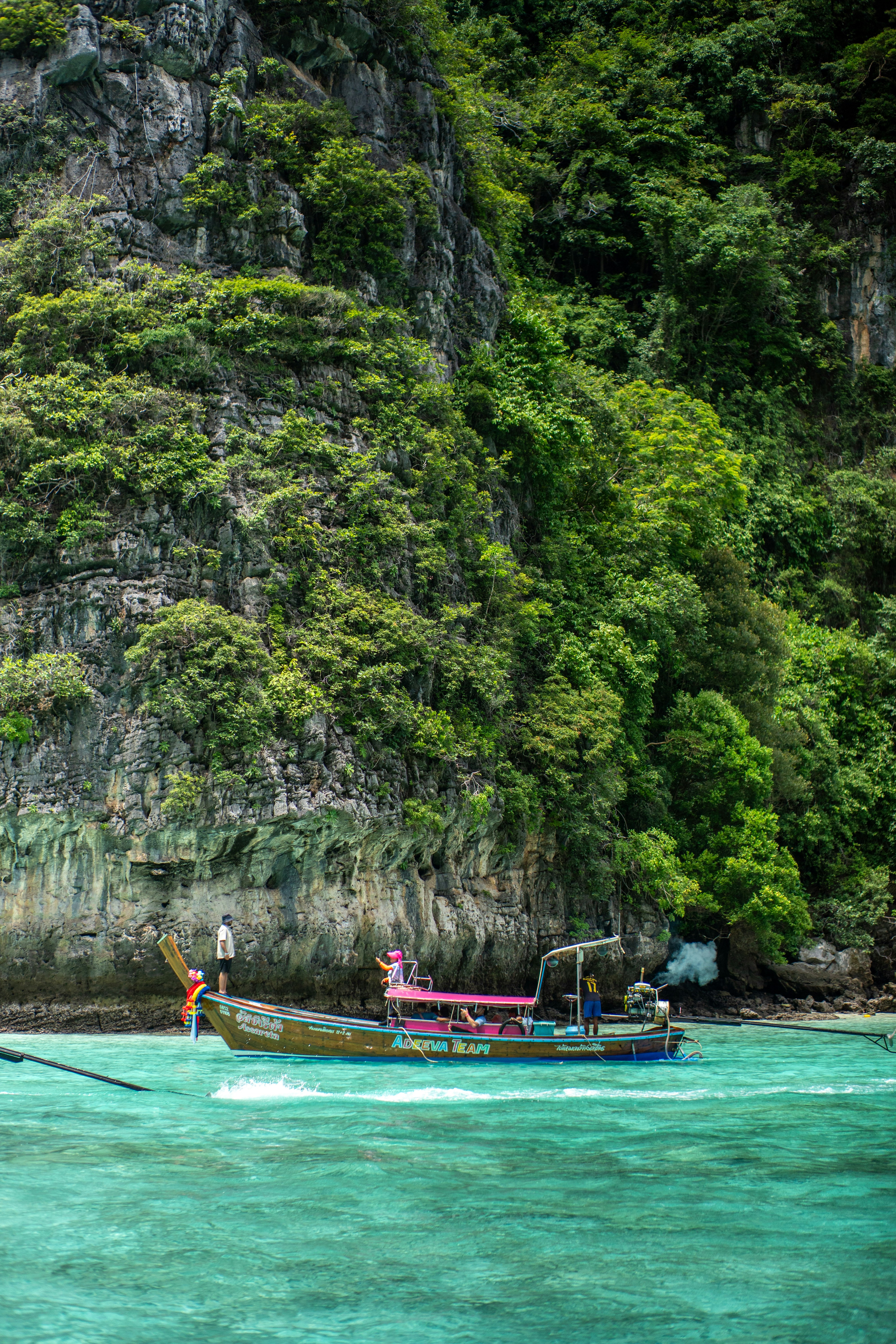 A longtail boat with people on turquoise water below a steep limestone cliff