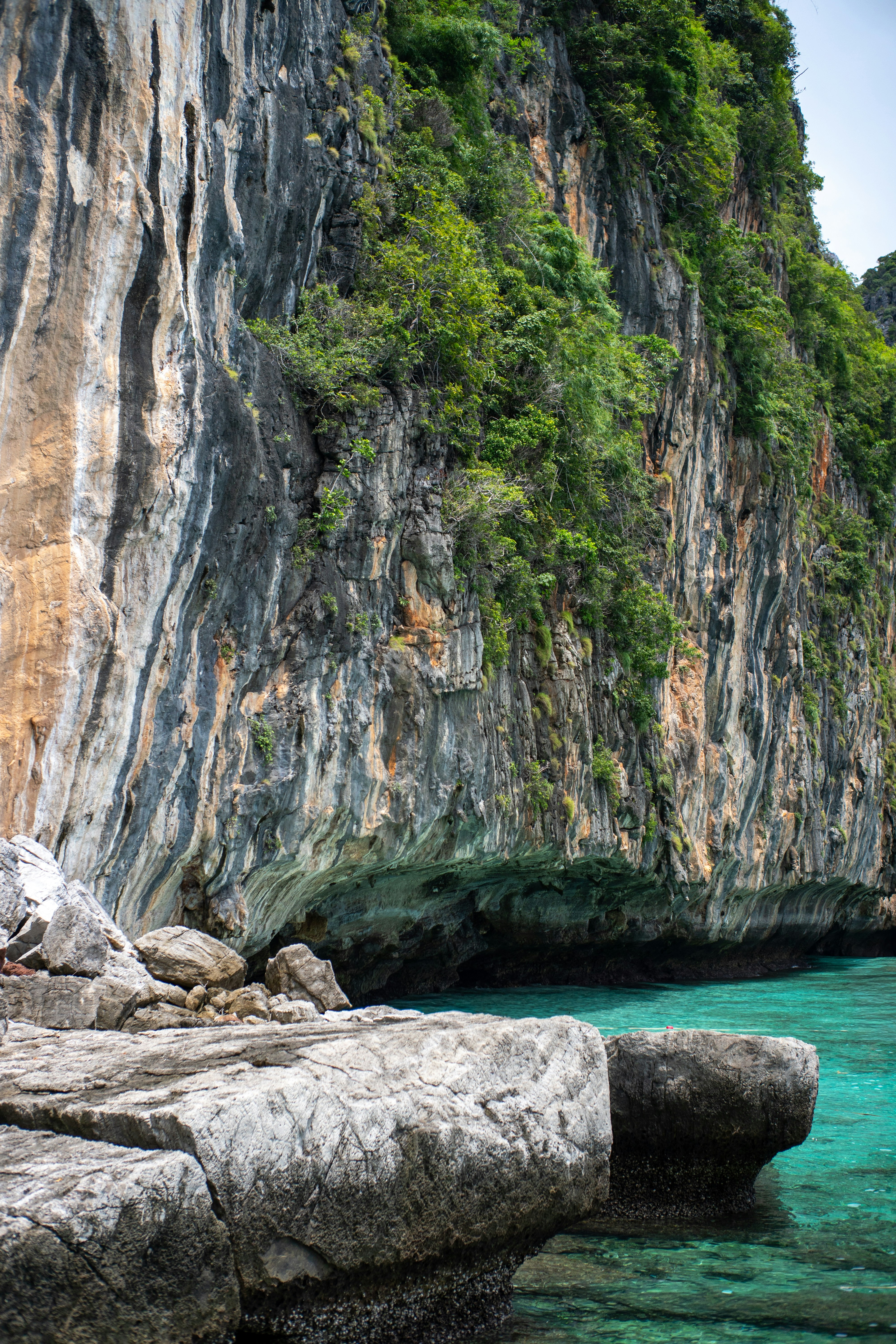 A towering limestone cliff with green vegetation above turquoise water and rocky boulders