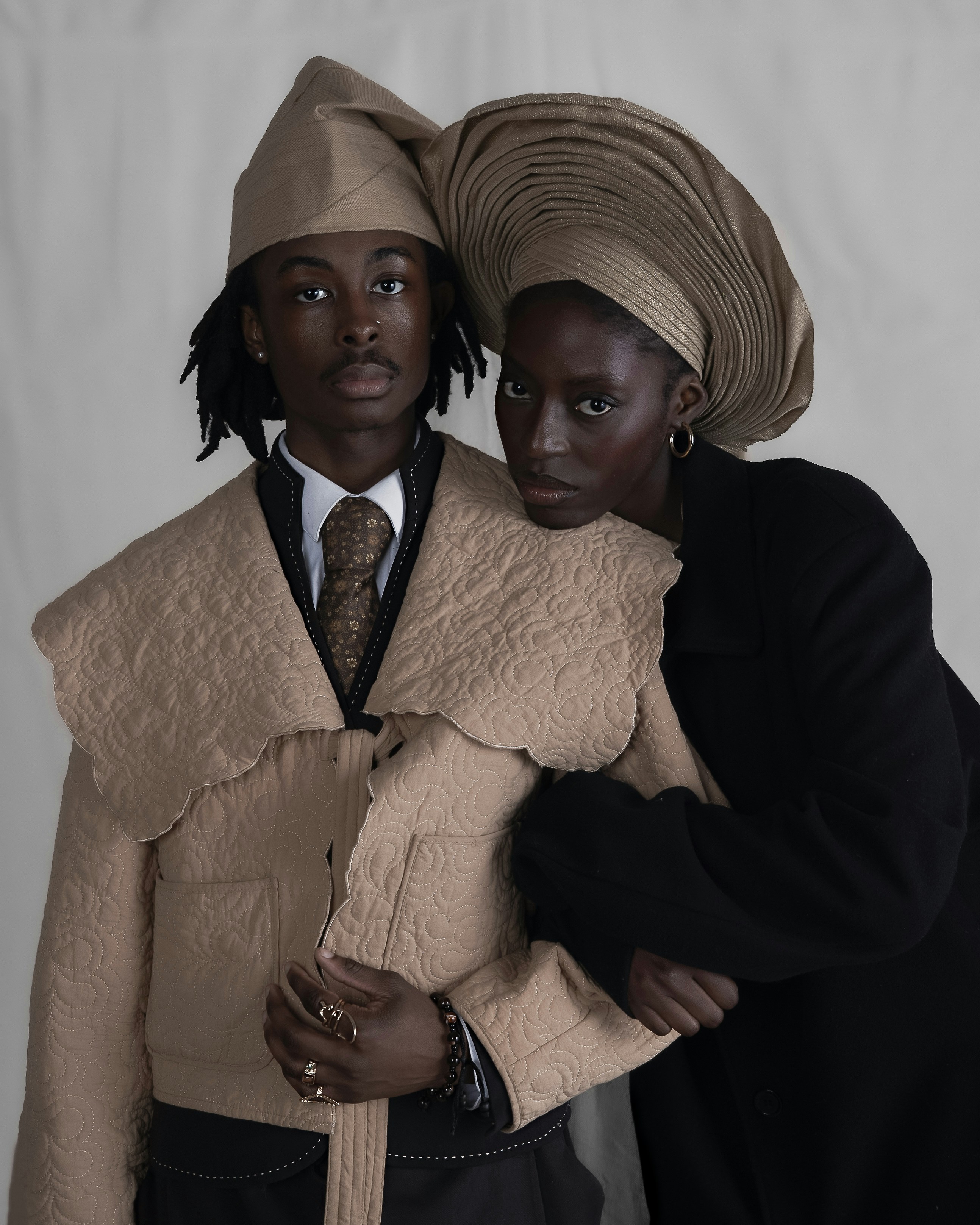 Two people in formal attire, one wearing a quilted beige jacket and headpiece