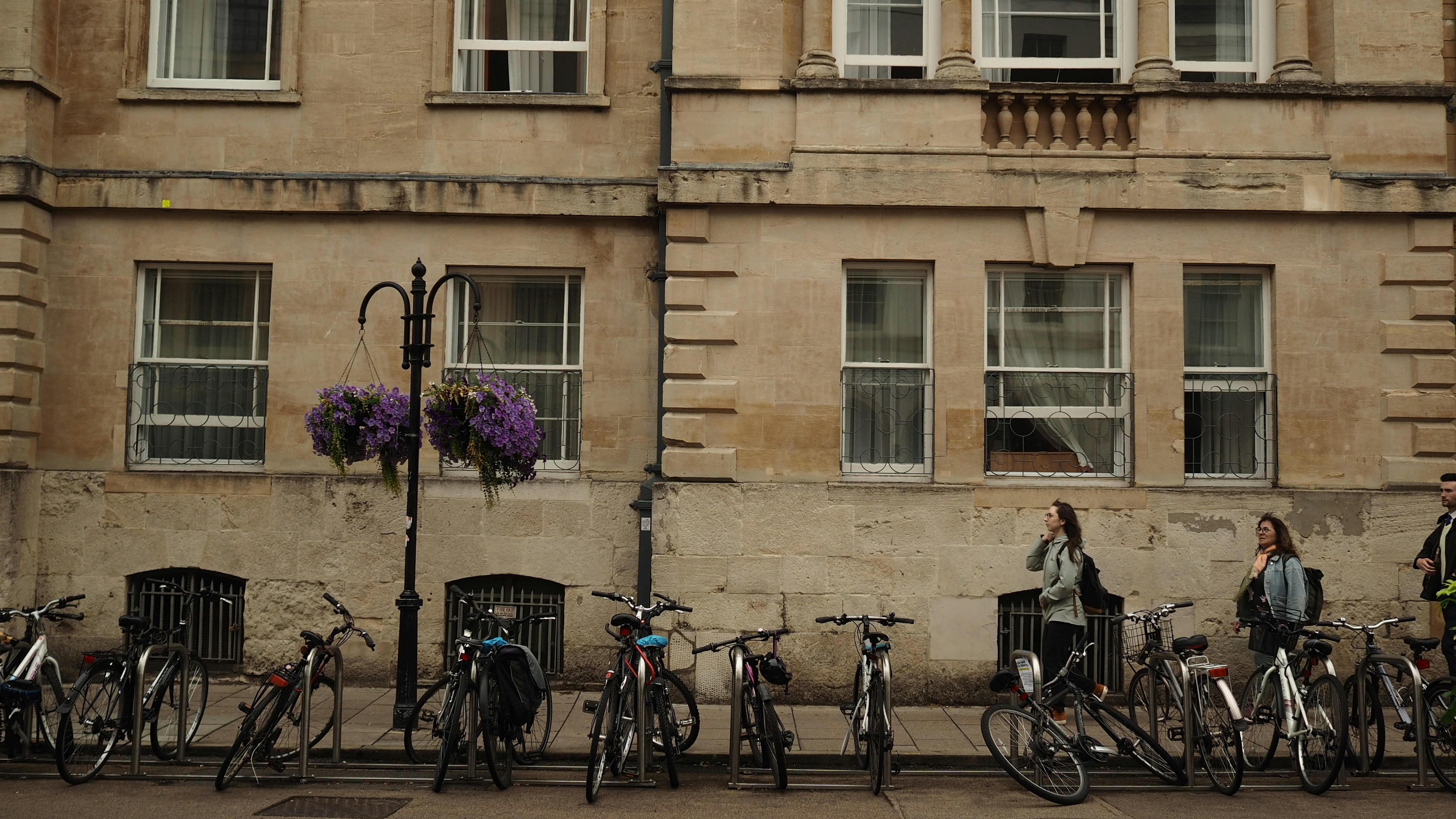 Des gens passant devant des vélos garés devant un bâtiment en pierre orné de fleurs suspendues